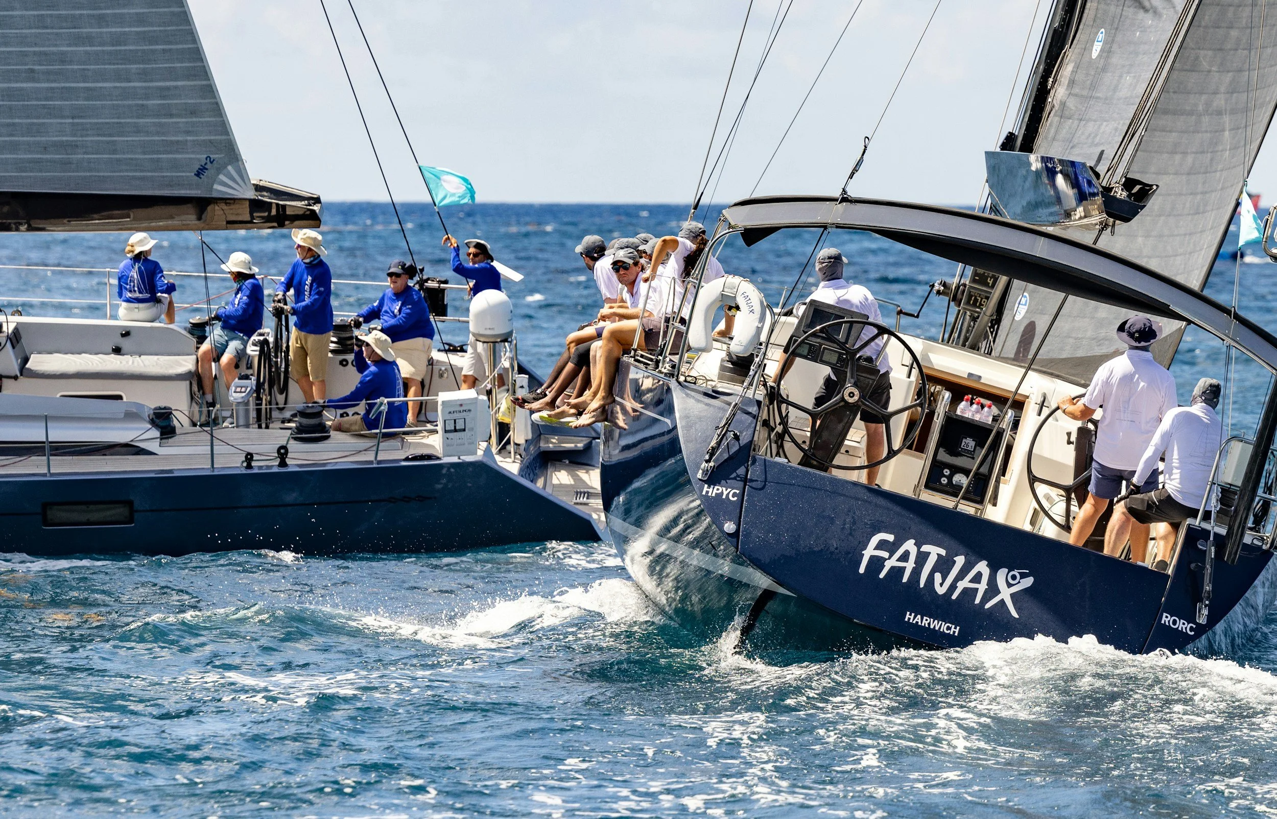 Två segelbåtar tävlar i en race på öppet hav med flera personer ombord, vissa som arbetar med seglen och andra som tittar och njuter av klimatet.