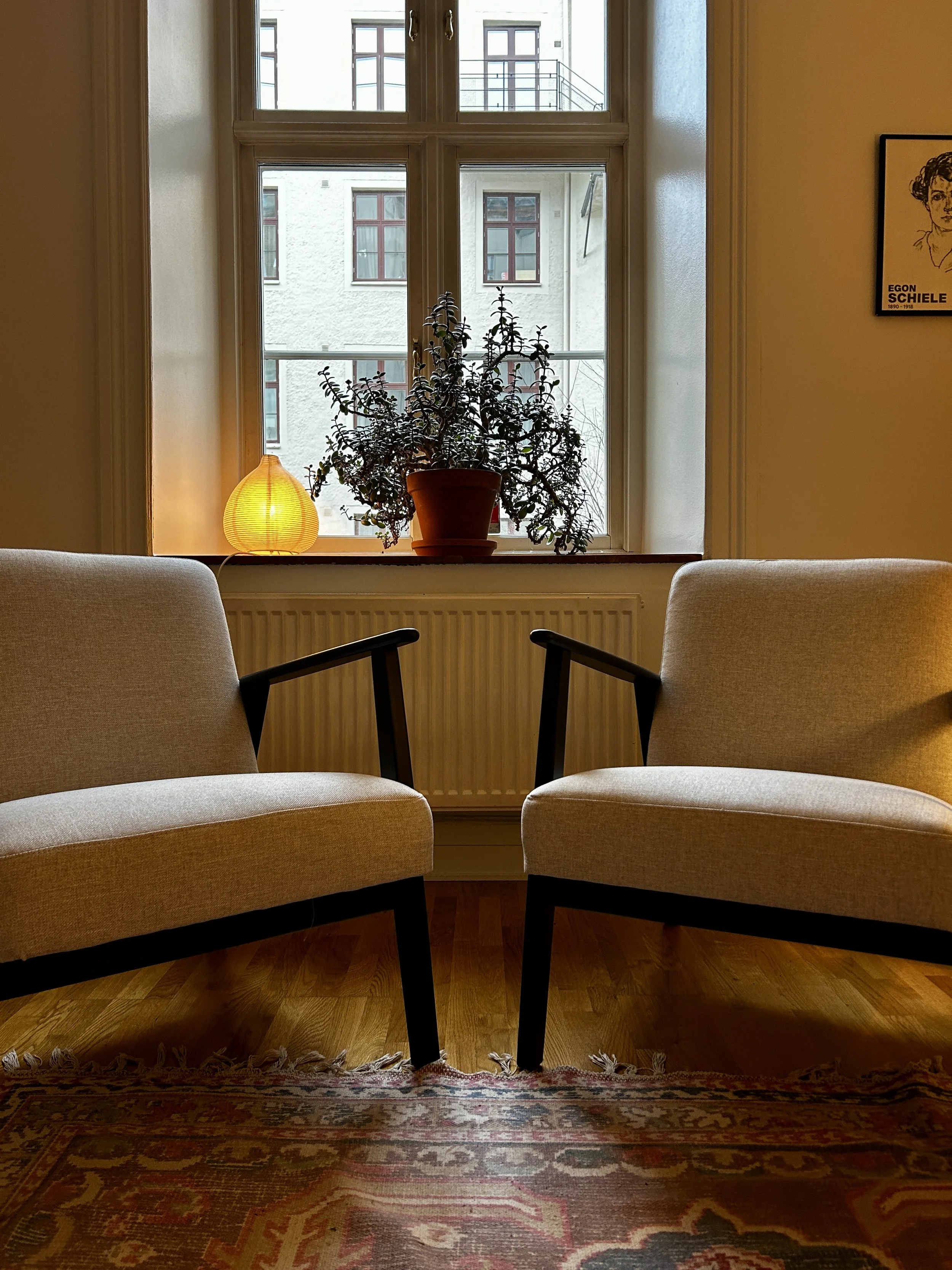 Två beige fåtöljer framför ett fönster med växter på fönsterbrädan, det finns en liten lampa och en tavla på väggen.