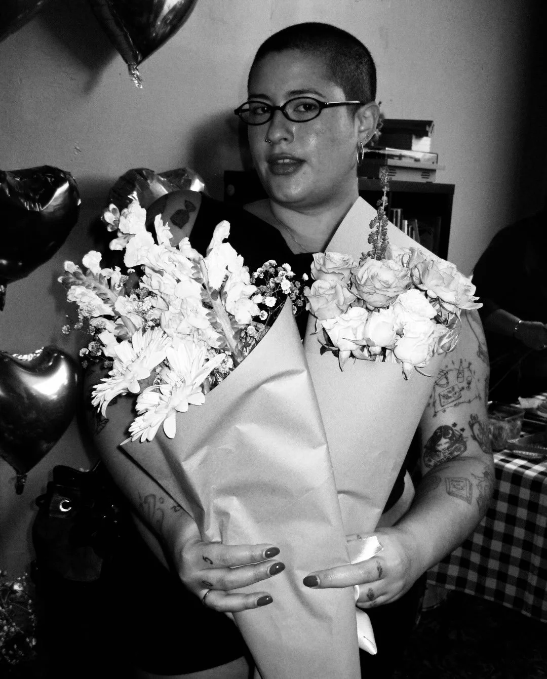 A person with short hair, glasses, and tattoos holding a large bouquet of flowers, surrounded by balloons, at an indoor celebration.