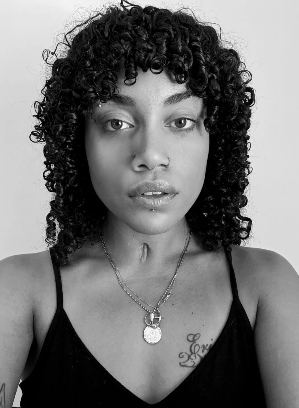 A black and white portrait of a woman with curly hair, facial piercings, and tattoos on her chest, wearing layered necklaces and a black spaghetti strap top.