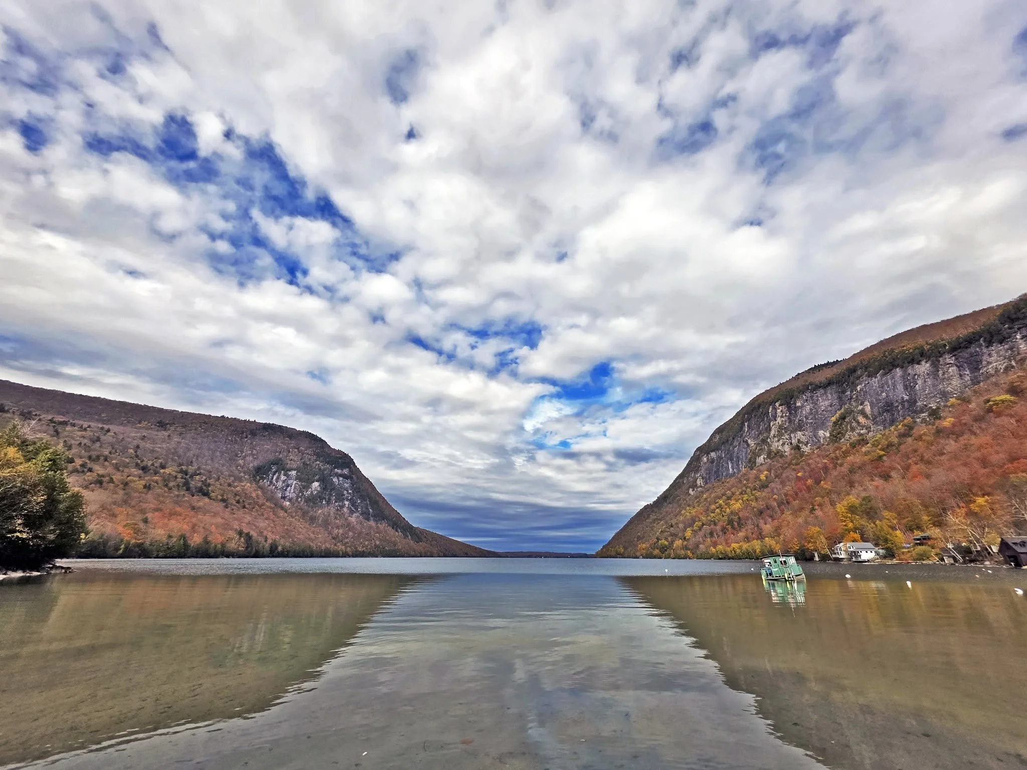 A serene lake surrounded by mountains with autumn foliage, under a partly cloudy sky.