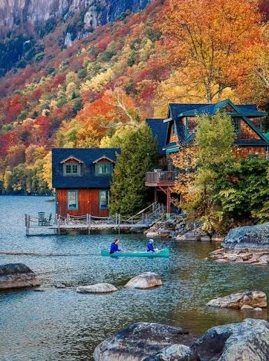 A lakeside scene with colorful autumn trees, two houses near the water, and two people in a kayak on the lake.