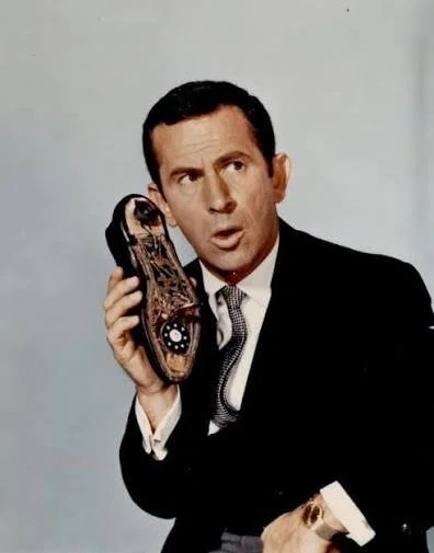 A man in a black suit holding a vintage rotary phone to his ear, with a surprised or confused expression.