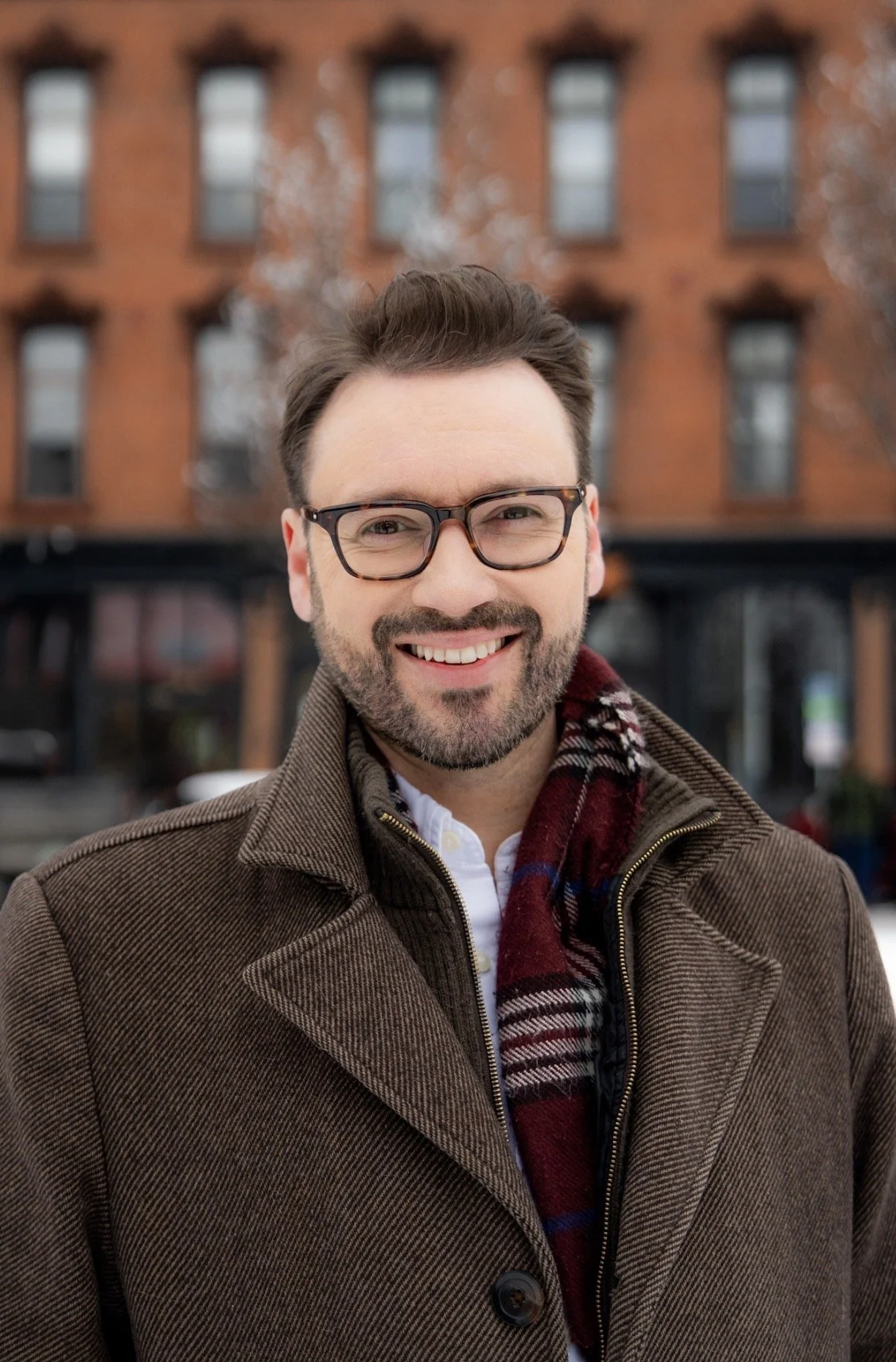 A smiling man with glasses and a beard wearing a brown coat and a plaid scarf standing outdoors in front of a brick building.
