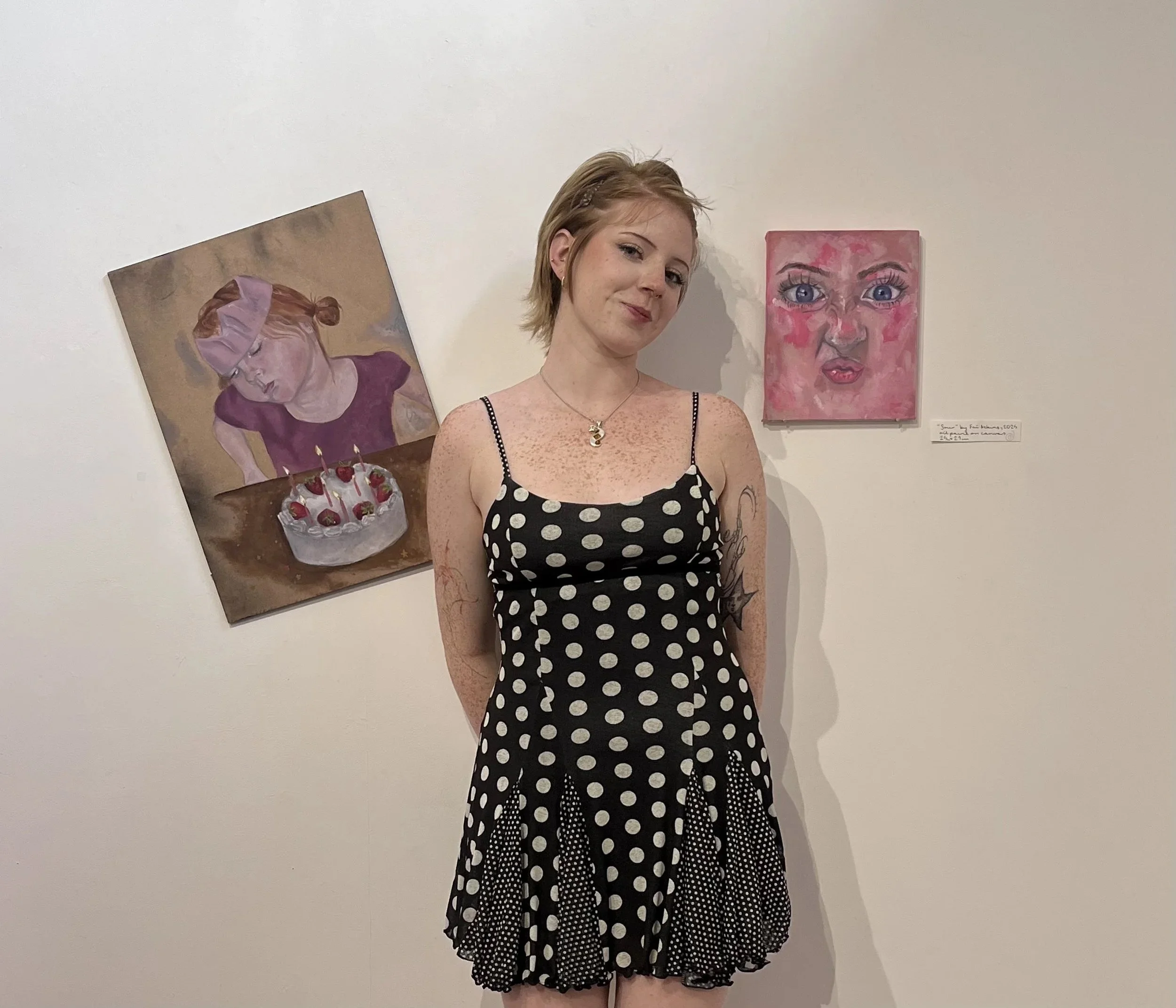 artist standing in front of two small scale paintings, one depicting herself as a child, the other depicting a close up portrait.