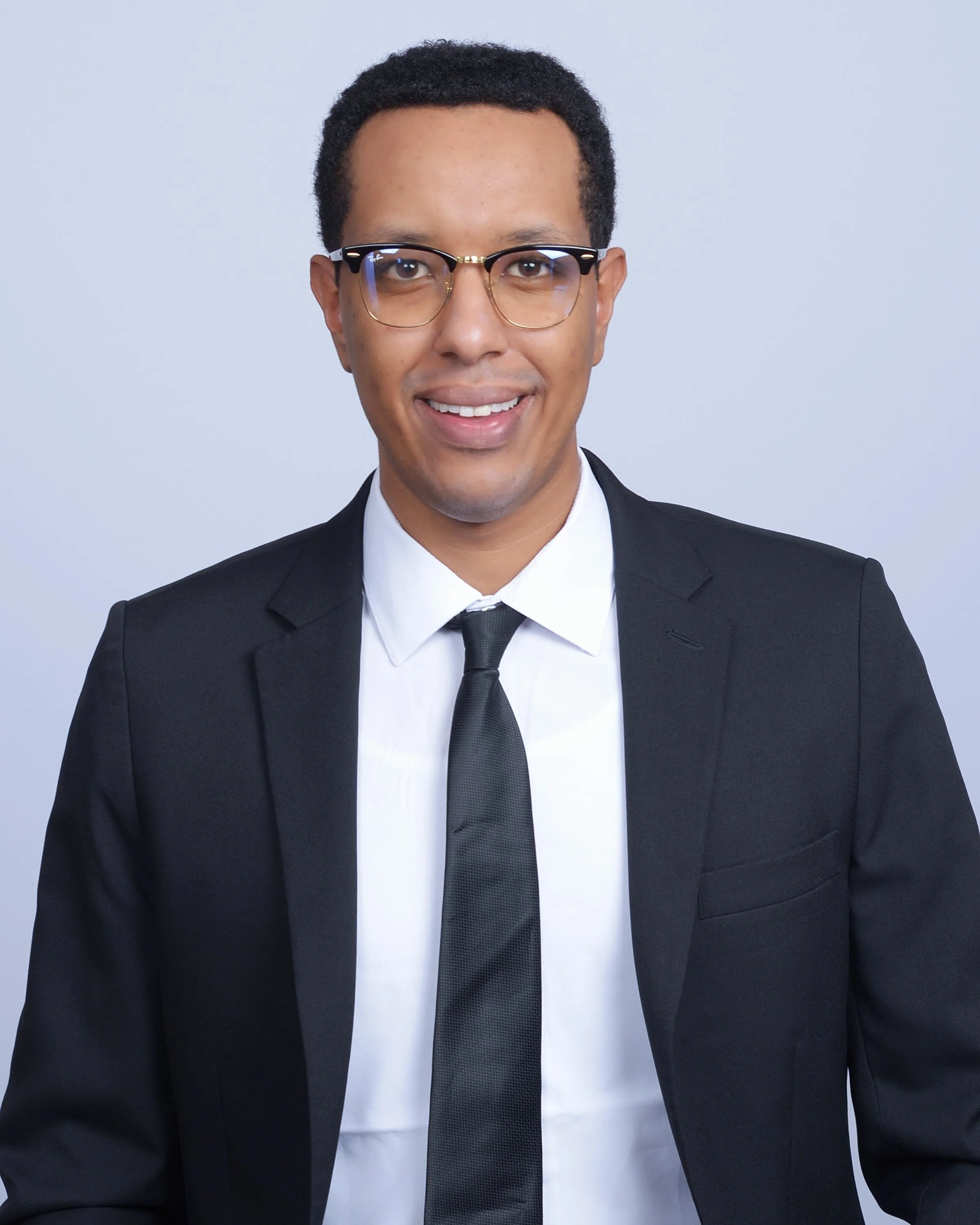 The man, CEO Abraham Teklu, wearing glasses, a white shirt, a black tie, and a black suit smiling against a light gray background.