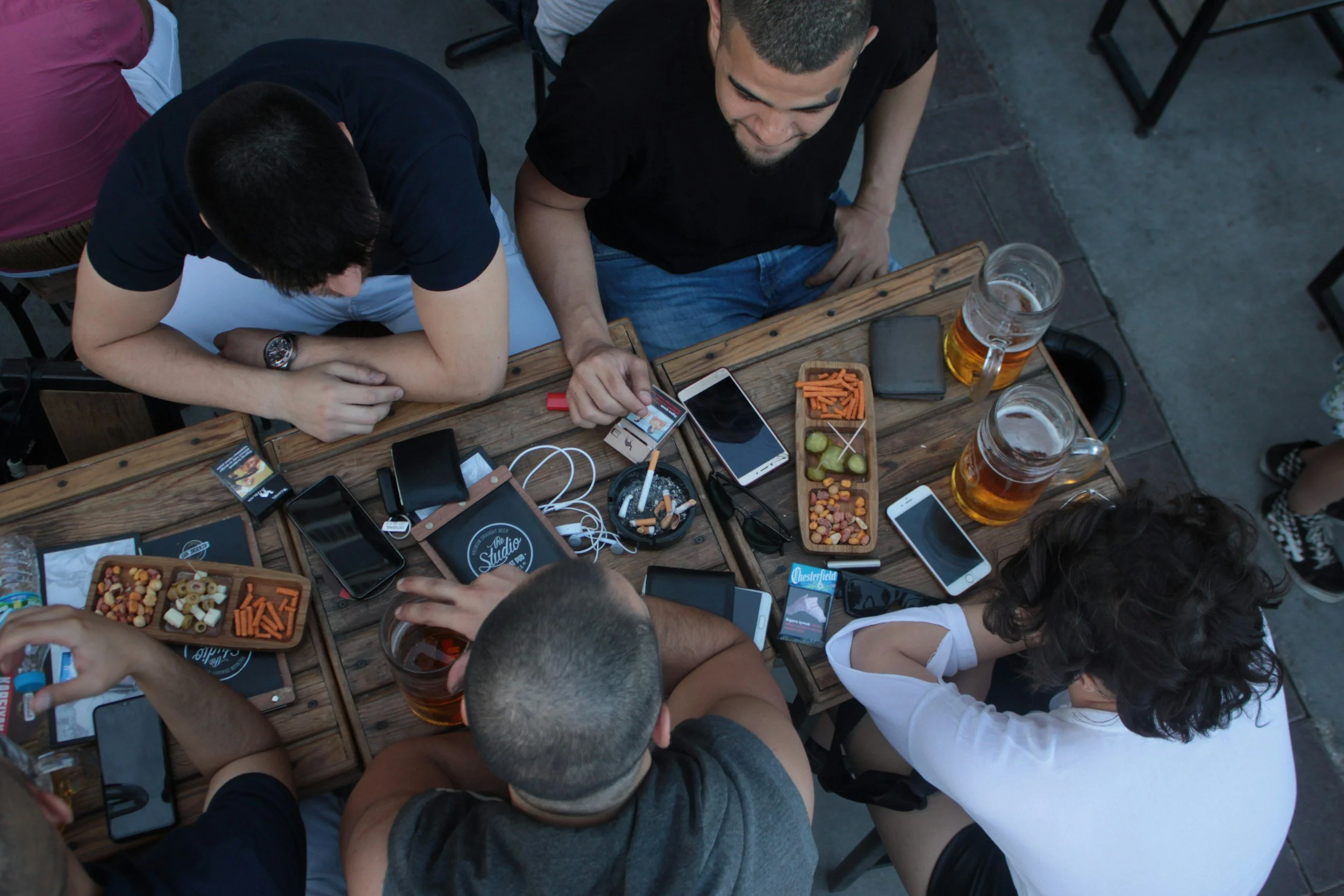 Group of people sitting around a wooden table at an outdoor gathering, with snacks and drinks, and using smartphones.