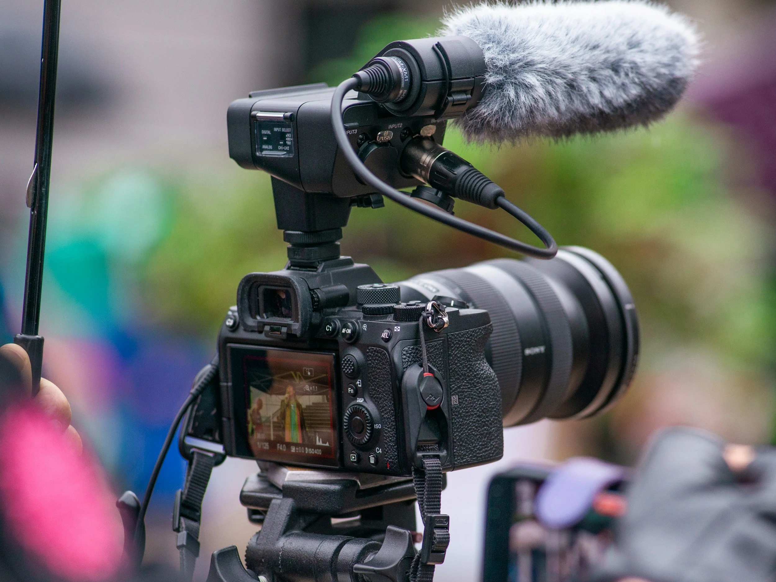 Professional video camera mounted on a tripod, with a large microphone with a furry windscreen attached on top, set outdoors with blurred background.