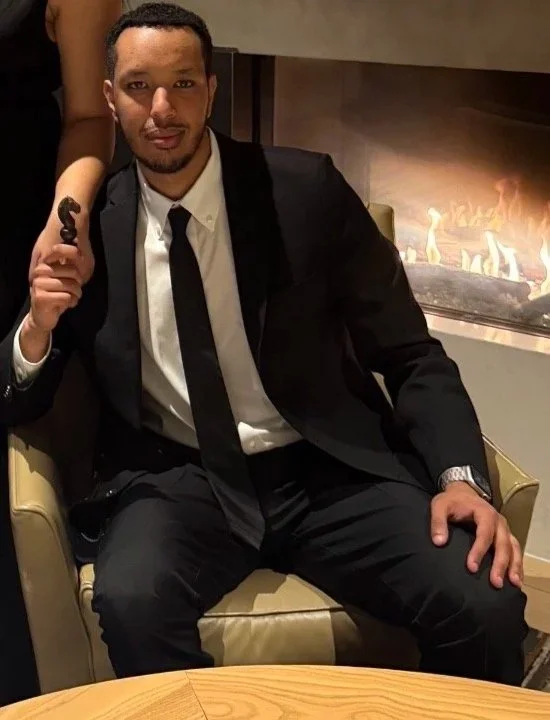 A man in a black suit, white shirt, and black tie sitting in a yellow armchair near a fireplace, holding a cigar in his right hand.