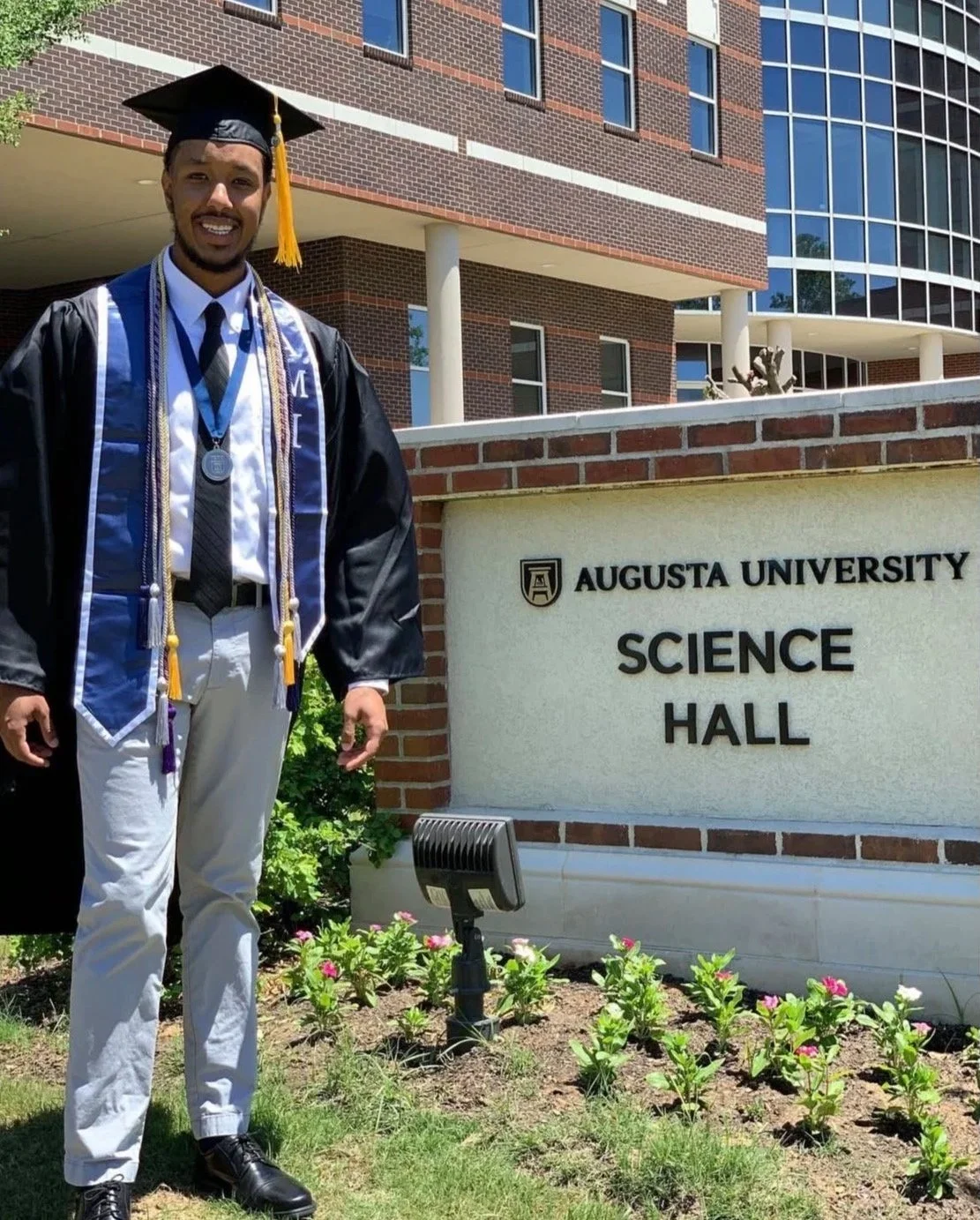 A young man in a graduation cap and gown standing outside Augusta University's Science Hall, smiling, wearing a medal and cords, with a brick building and flowers in the background.