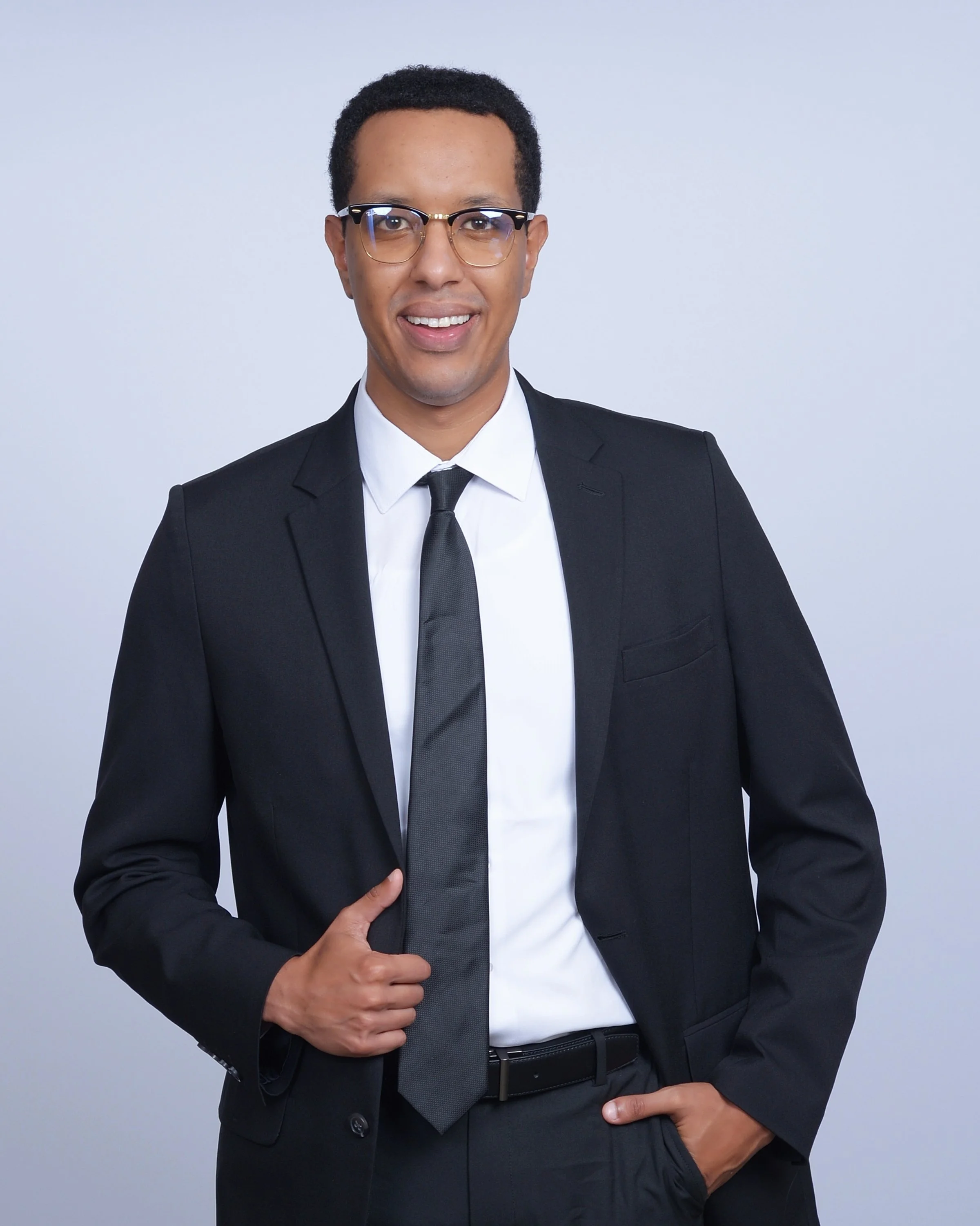 A man in a black suit, white dress shirt, and black tie smiling with glasses against a gray background.