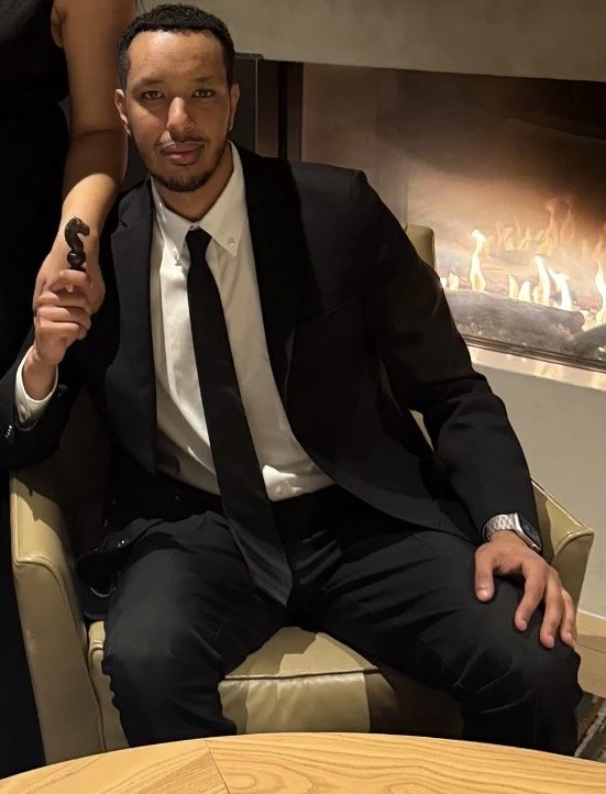 The man, CEO Abraham Teklu, in a black suit, white shirt, and black tie sitting in a beige chair next to a fireplace, holding a small chess piece in his right hand.