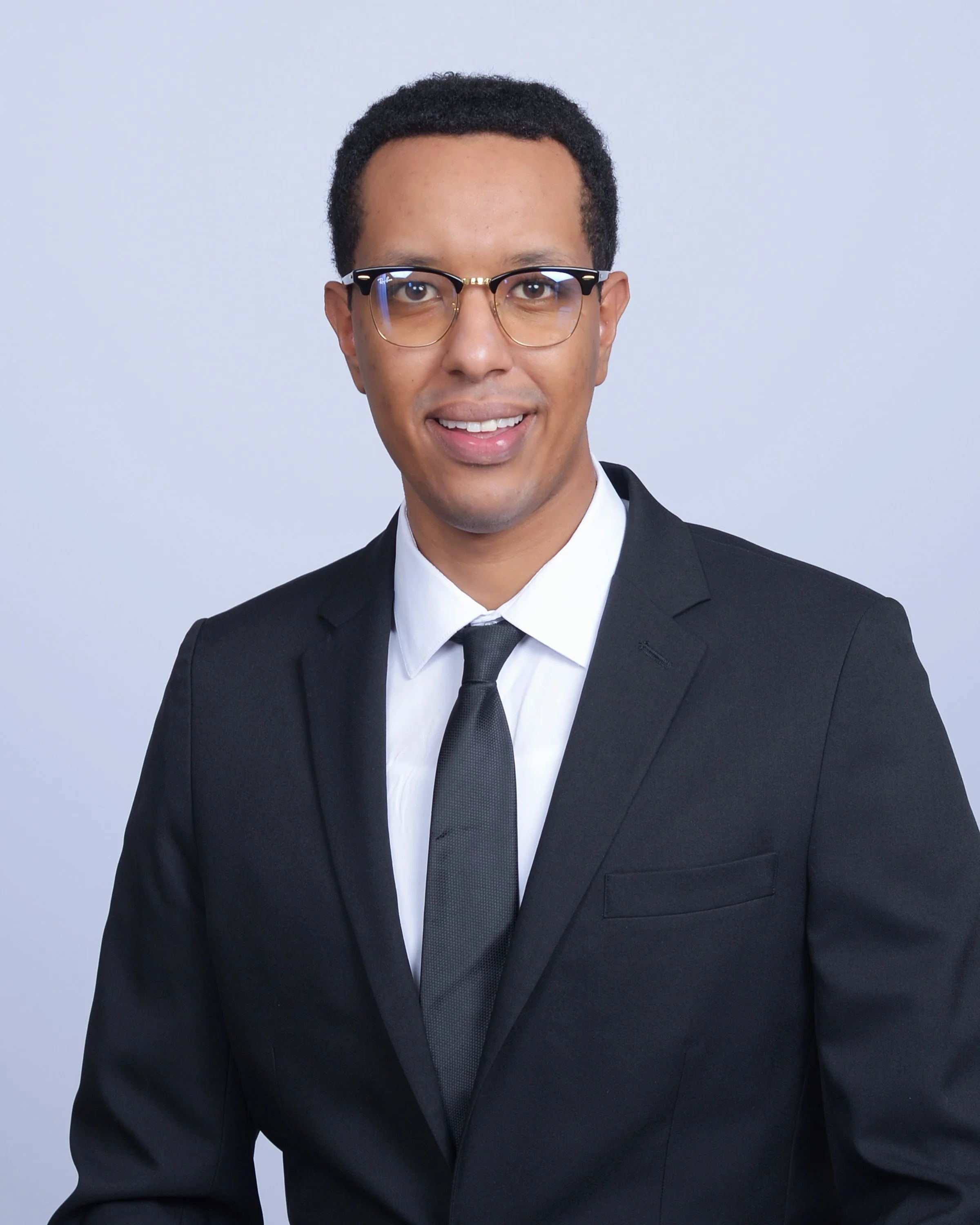 The man, CEO Abraham Teklu, in a black suit, white shirt, and black tie is smiling and wearing glasses with a light background.