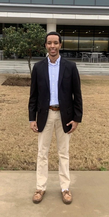A man in business casual attire standing outdoors in front of a modern building with glass windows, trees, and outdoor seating.