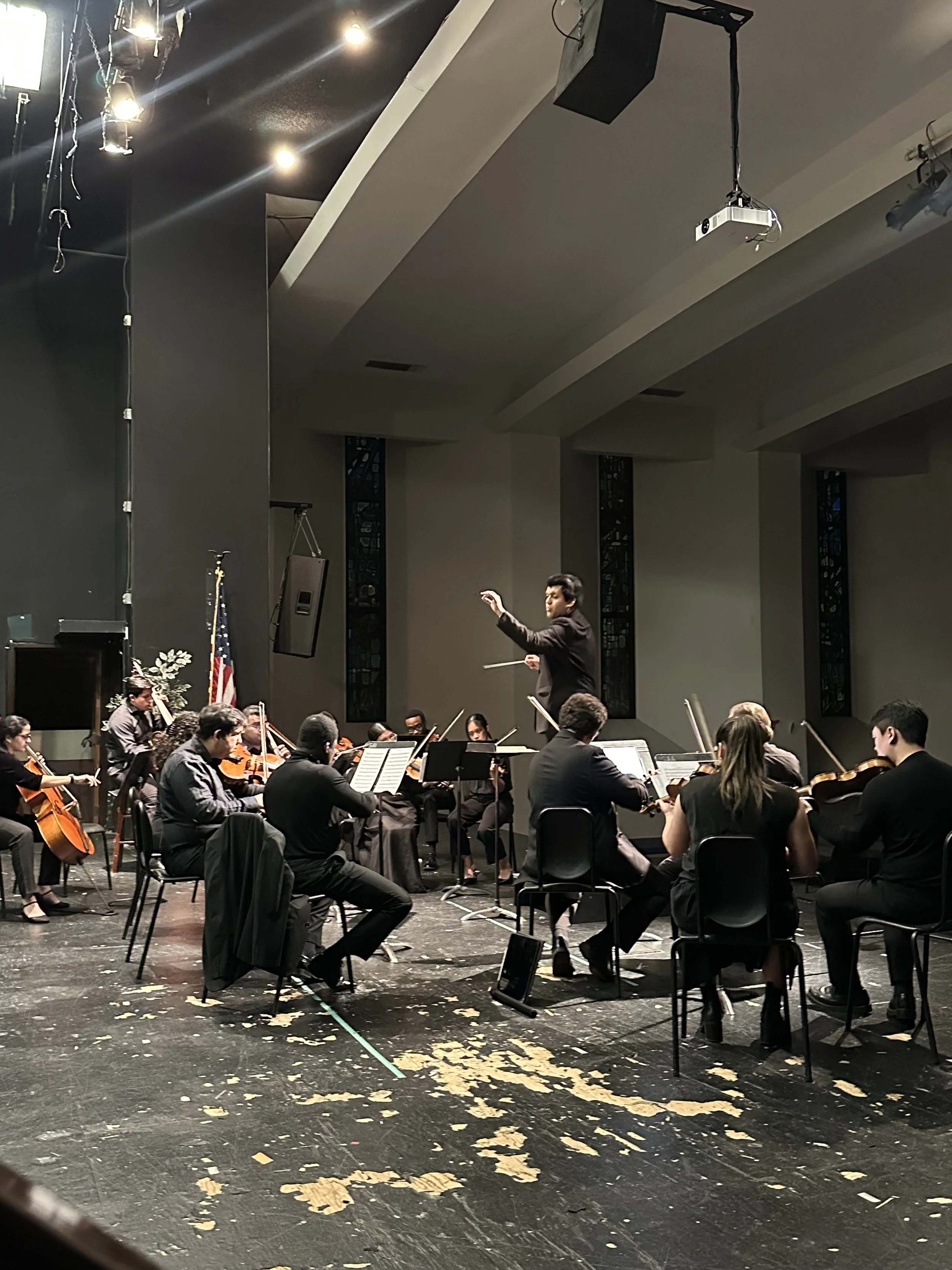 An orchestra performing on stage under conductor's direction, with violins and cellos, in a concert hall with American flag and stained glass windows.