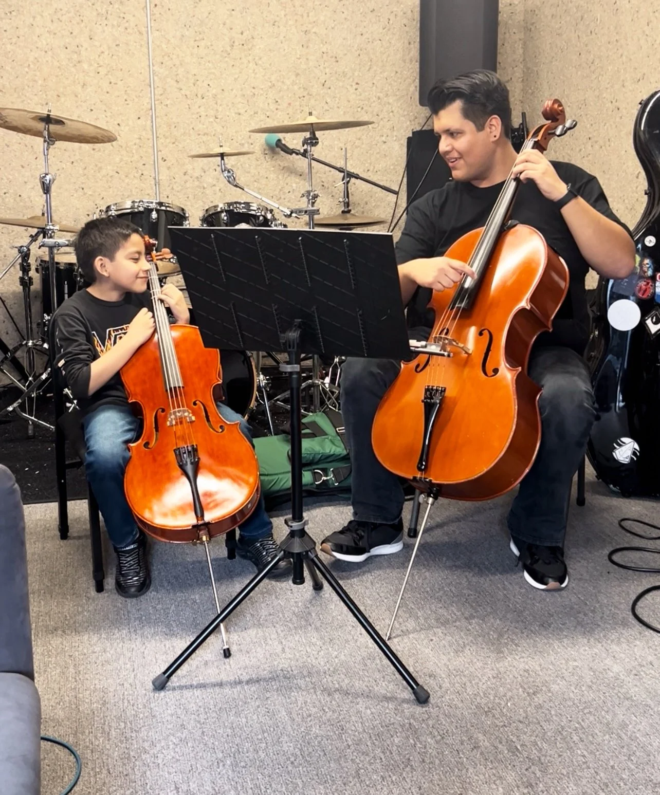 Adult male teaching a young student how to play cello. The teacher looks like he enjoys teaching and the young student seems to be having fun while playing and learning.