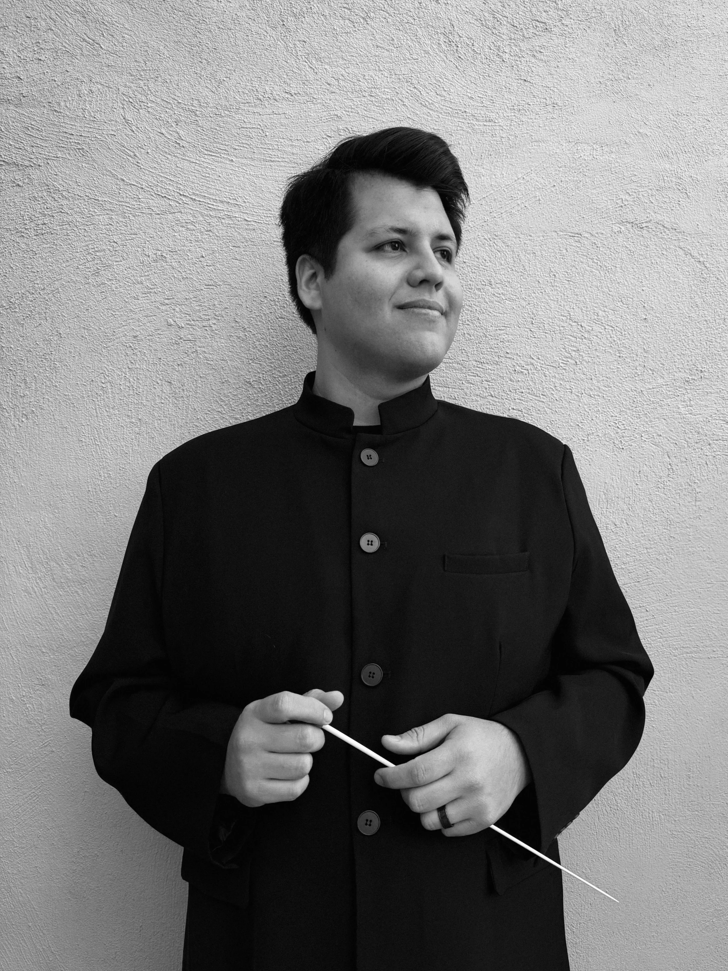 A person with short dark hair wearing a traditional Bangladeshi outfit standing against a plain textured wall, holding a baton.