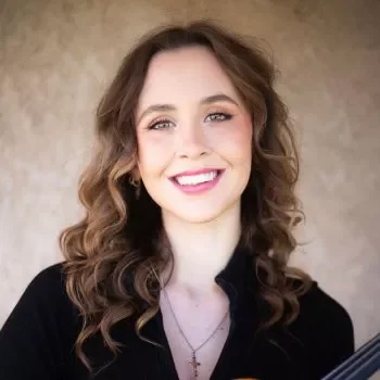 A young woman with curly brown hair smiling at the camera, wearing a black top and standing against a neutral background.