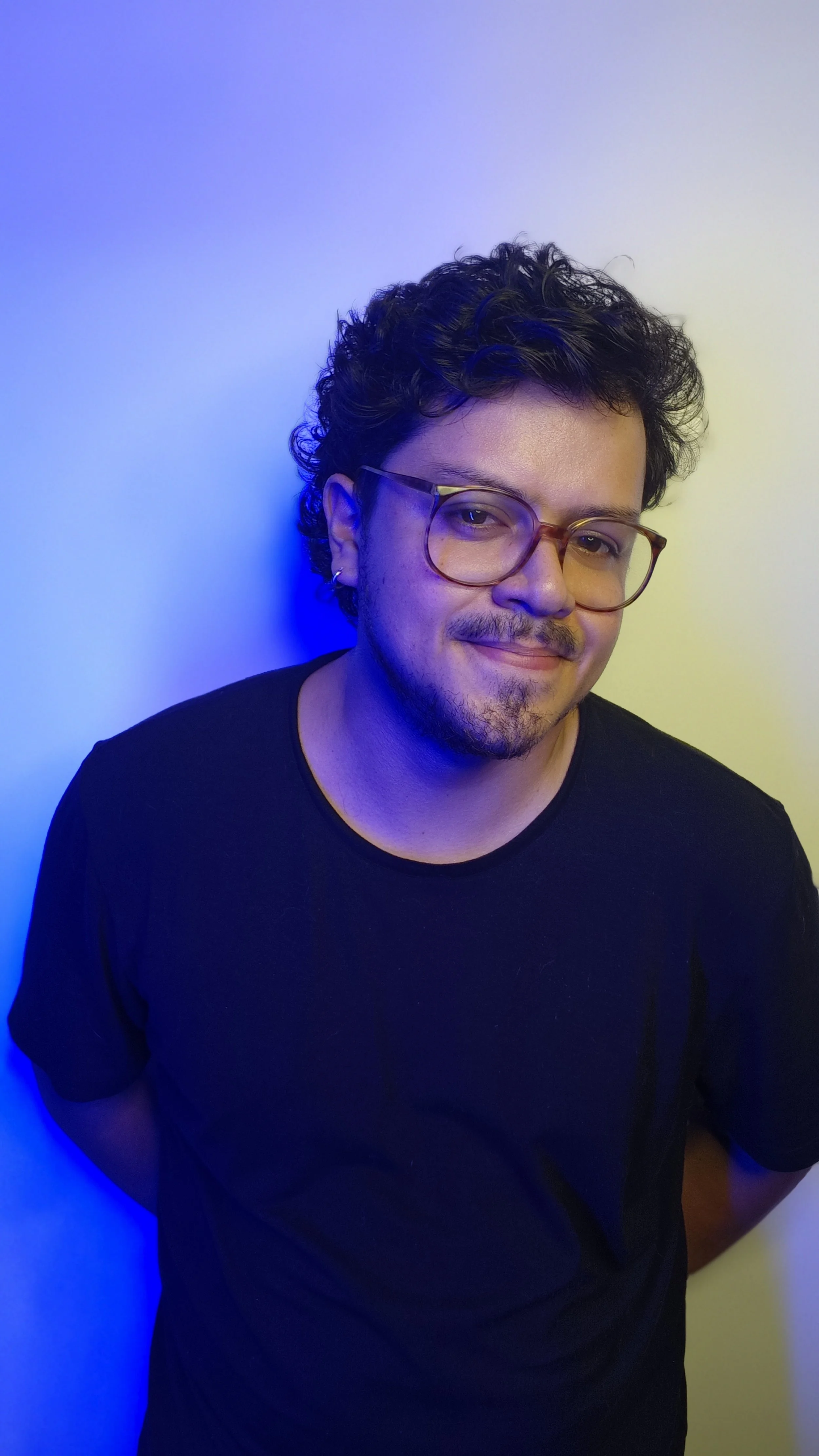 Joven con gafas, cabello rizado, sonriendo, de fondo de paredes azul y blanca con luces de colores.