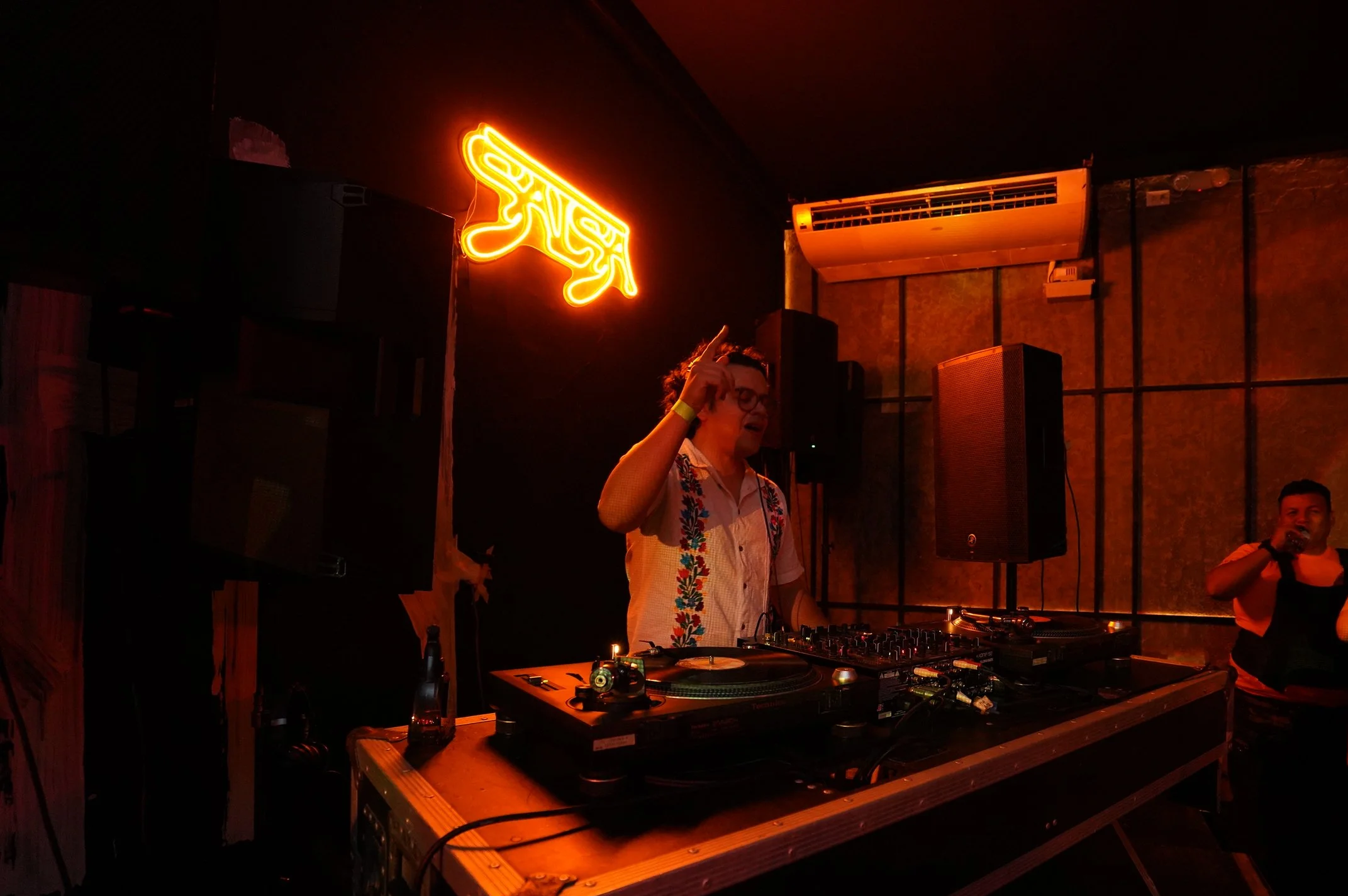 Un DJ tocando en un establecimiento con decoración de neón y una persona al fondo.