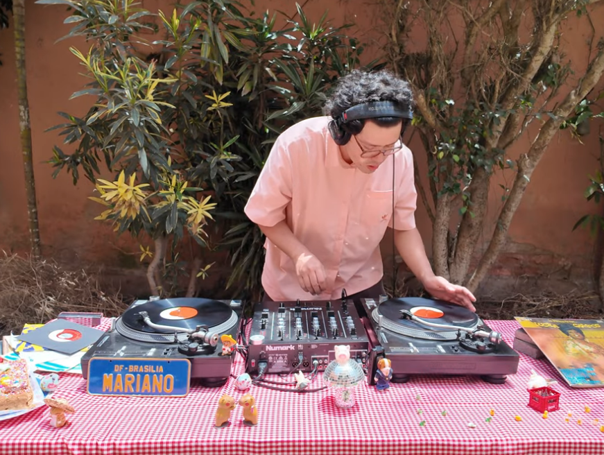 Una persona mezclada en la música usando audífonos, operando un DJ, en una mesa decorada con juguetes y otros objetos, con plantas y árboles en el fondo.