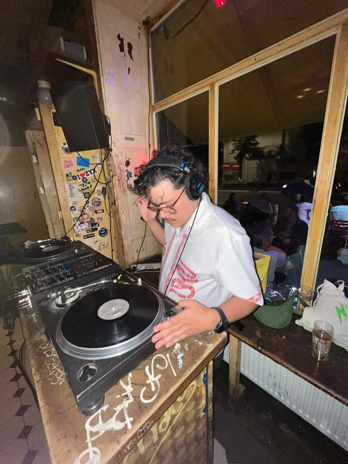 Un DJ con auriculares operando una tornamesa en un ambiente con una gran ventana, con varias personas en el fondo y decoraciones en la pared.