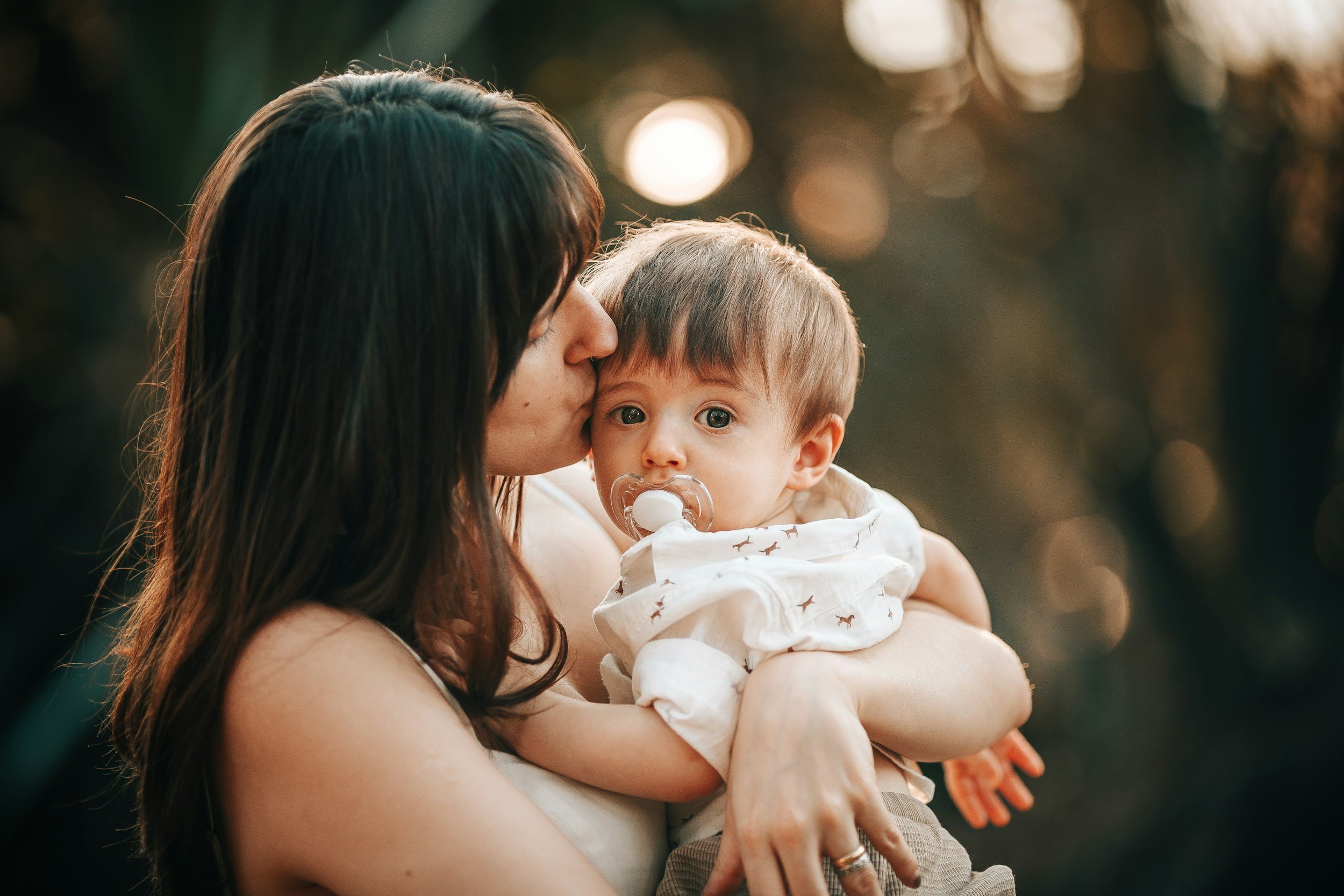 A woman holding a young child with a pacifier, kissing the child on the cheek outdoors during sunset.