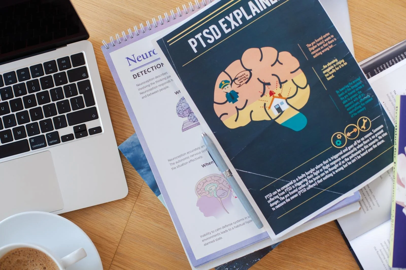 An open booklet on PTSD explanations with an illustration of a brain and house, surrounded by a police officer figure. A pen and papers are on top of the booklet, with a laptop and a cup of coffee nearby on a wooden desk.