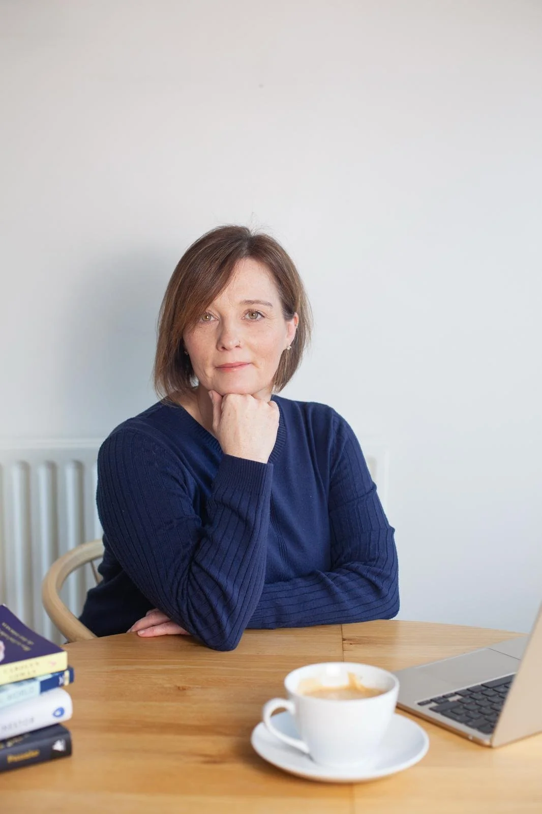 A woman with short brown hair wearing a blue sweater, sitting at a wooden table with a white cup of coffee, a laptop, and a few books.