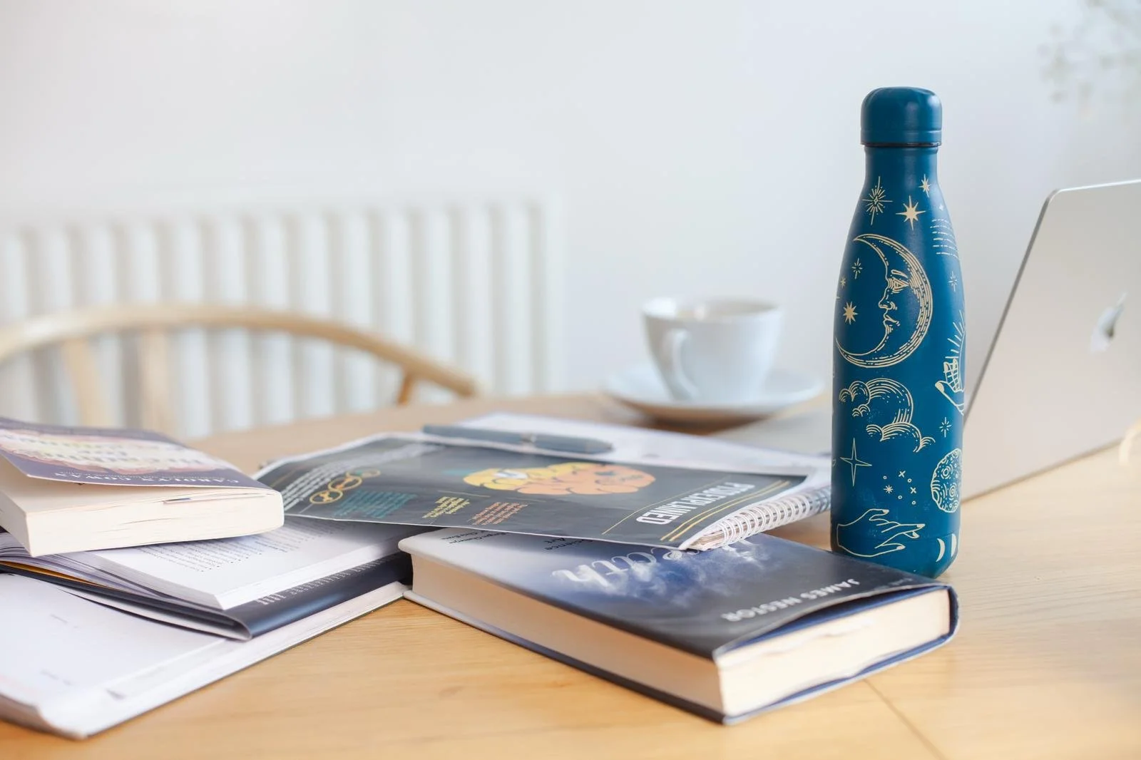 Messy workspace on a wooden table with notebooks, magazines, an astrology water bottle with moon and star designs, a teacup, and a laptop in a well-lit room.