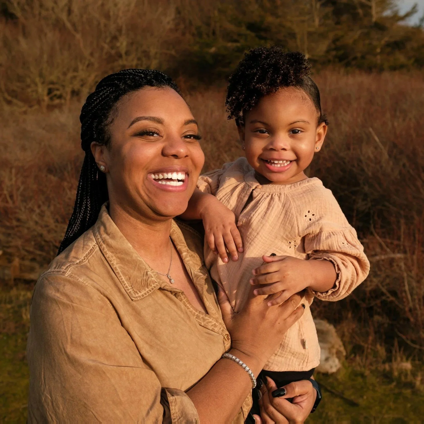 A smiling woman holding a happy young girl outdoors during sunset, with trees and dry grass in the background.