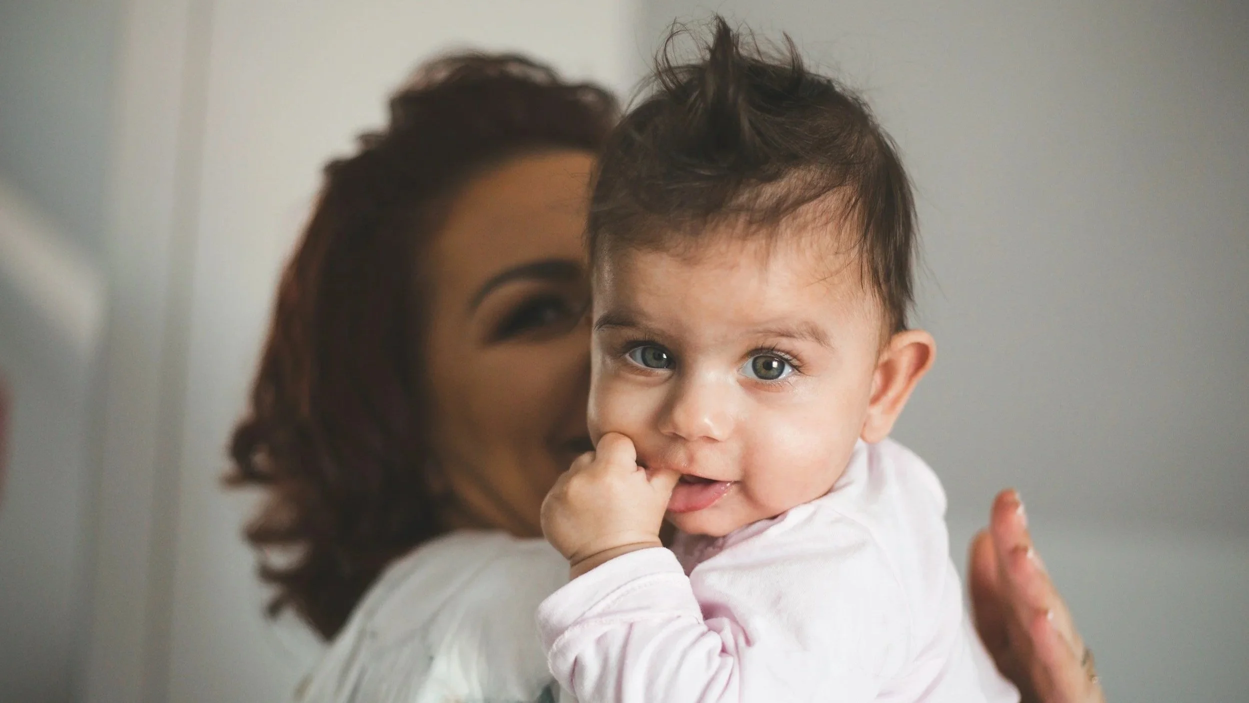 A woman holding a young child with blue eyes and messy brown hair, looking at the camera with the child sucking on their finger.