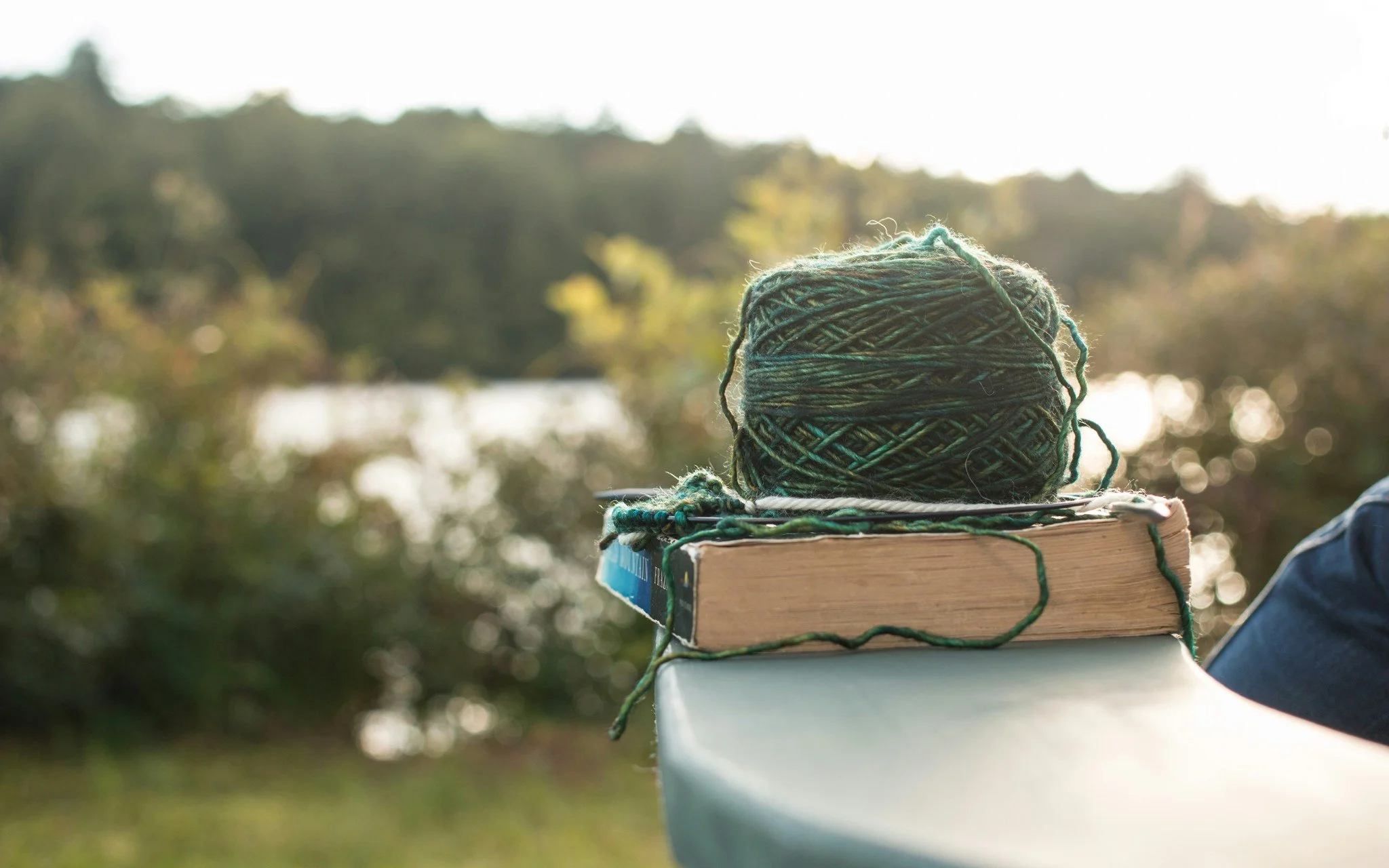 A ball of multicolored yarn resting on a closed book, placed on a smooth outdoor surface against a backdrop of trees and sunlight.