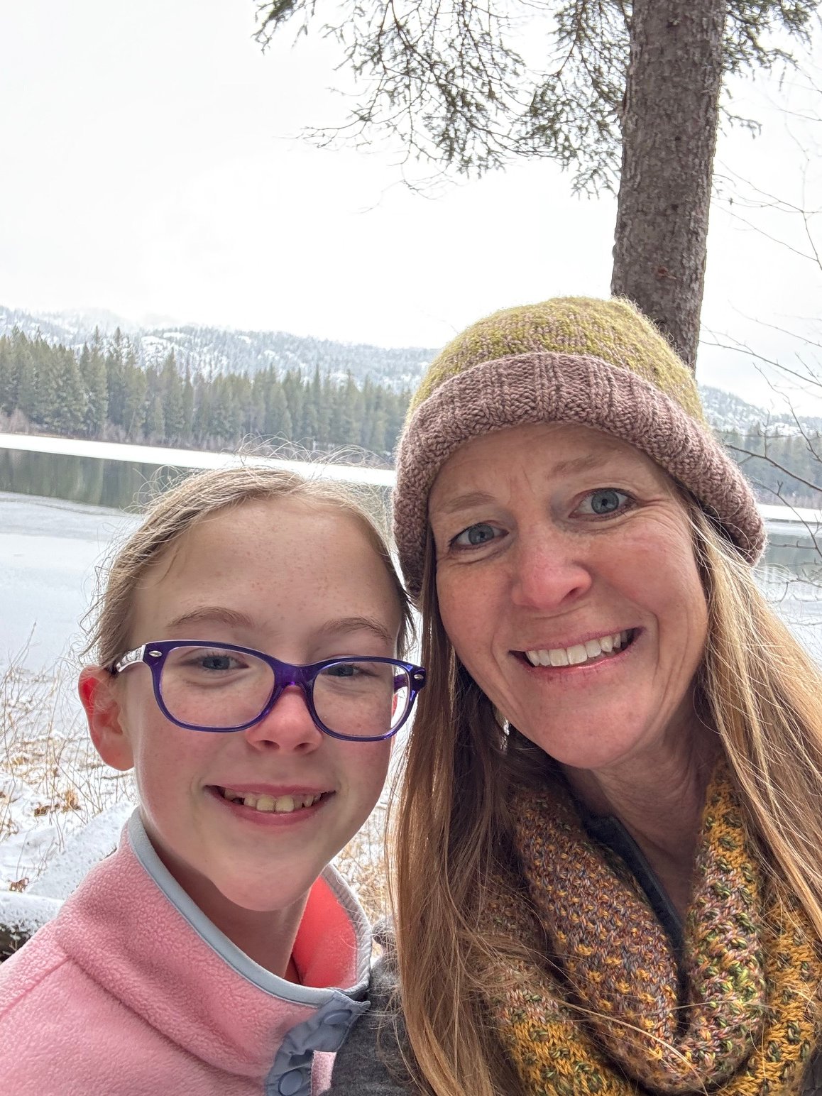 A mother and daughter team, inspiring knitwear, smiling for a selfie outdoors near a river with evergreen trees and snow-covered mountains in the background, one wearing a knit hat and the other glasses.