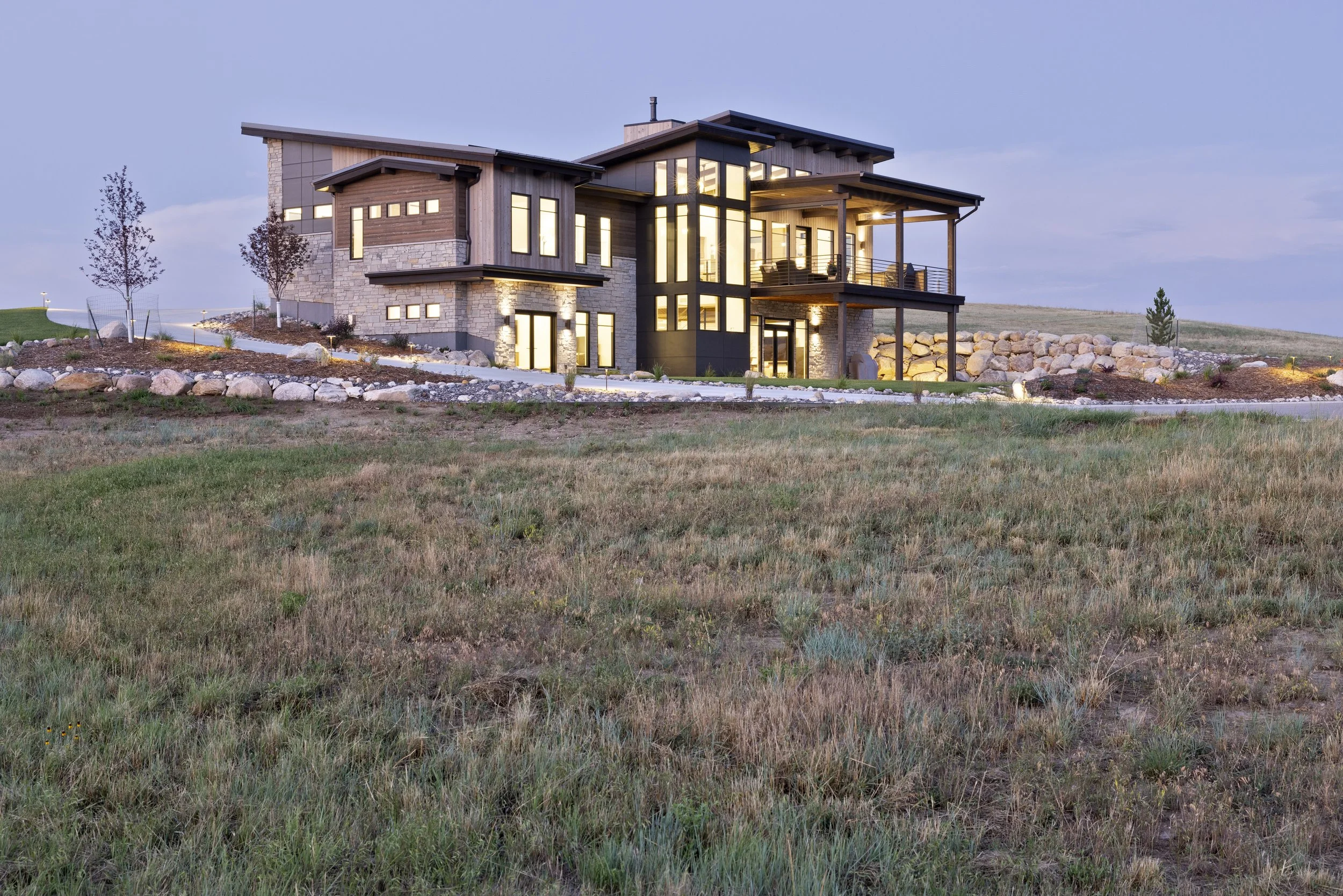 Modern house with large glass windows and a balcony, illuminated from inside, surrounded by a landscaped yard with trees and rocks.