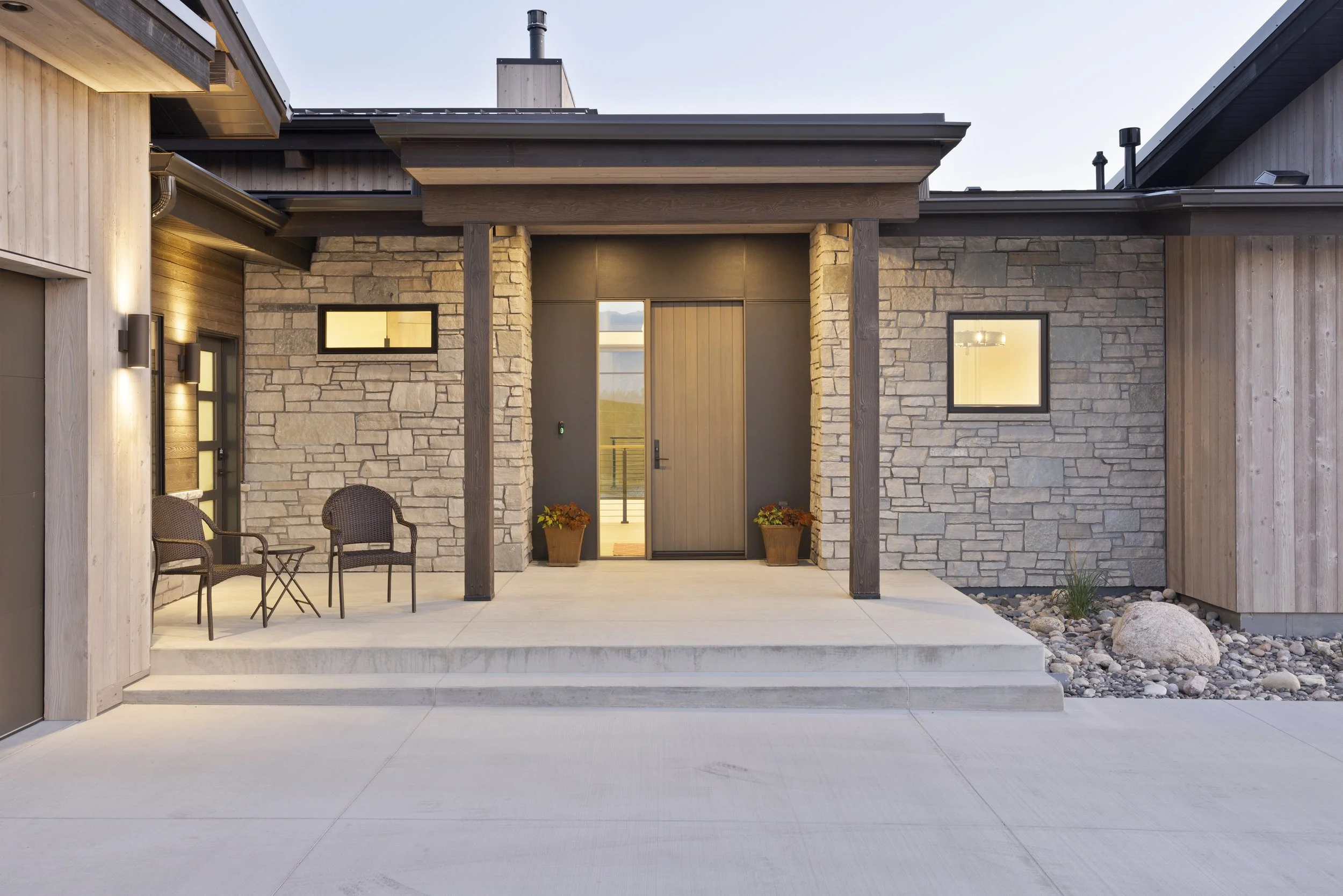Front view of a modern house entrance with stone and wood exterior, two small potted plants, outdoor seating, and large concrete patio.