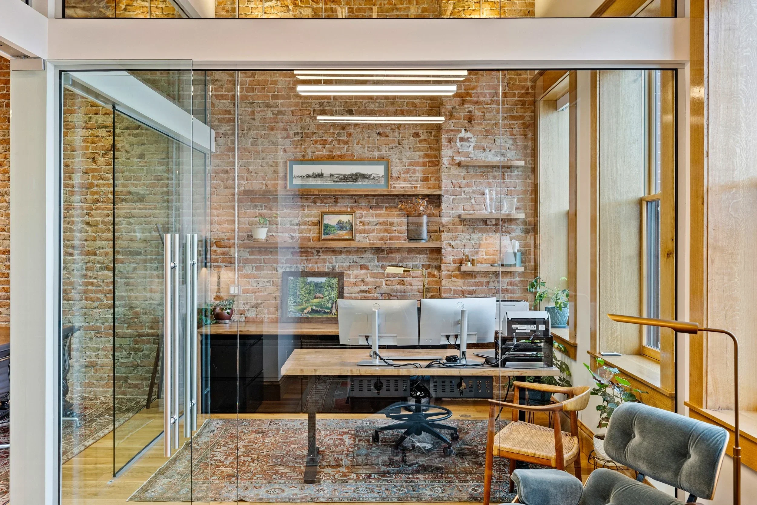 Modern office with brick walls, glass walls, and large windows, featuring a work desk with two computers, wall art, houseplants, and furniture.