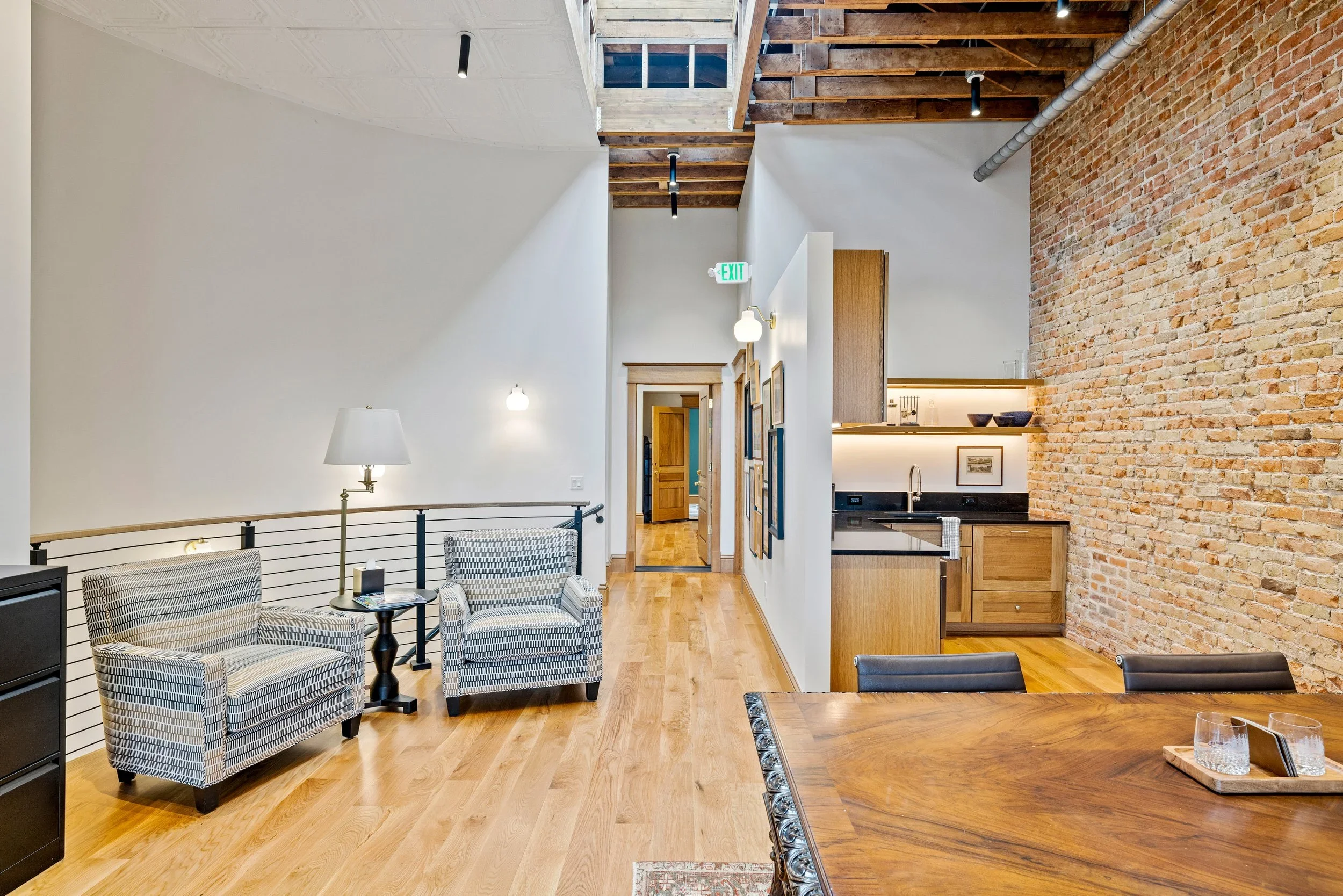 Interior of a modern apartment with hardwood floors, a brick accent wall, a kitchen with wooden cabinets and open shelves, and a sitting area with striped armchairs, a table, and standing lamps.