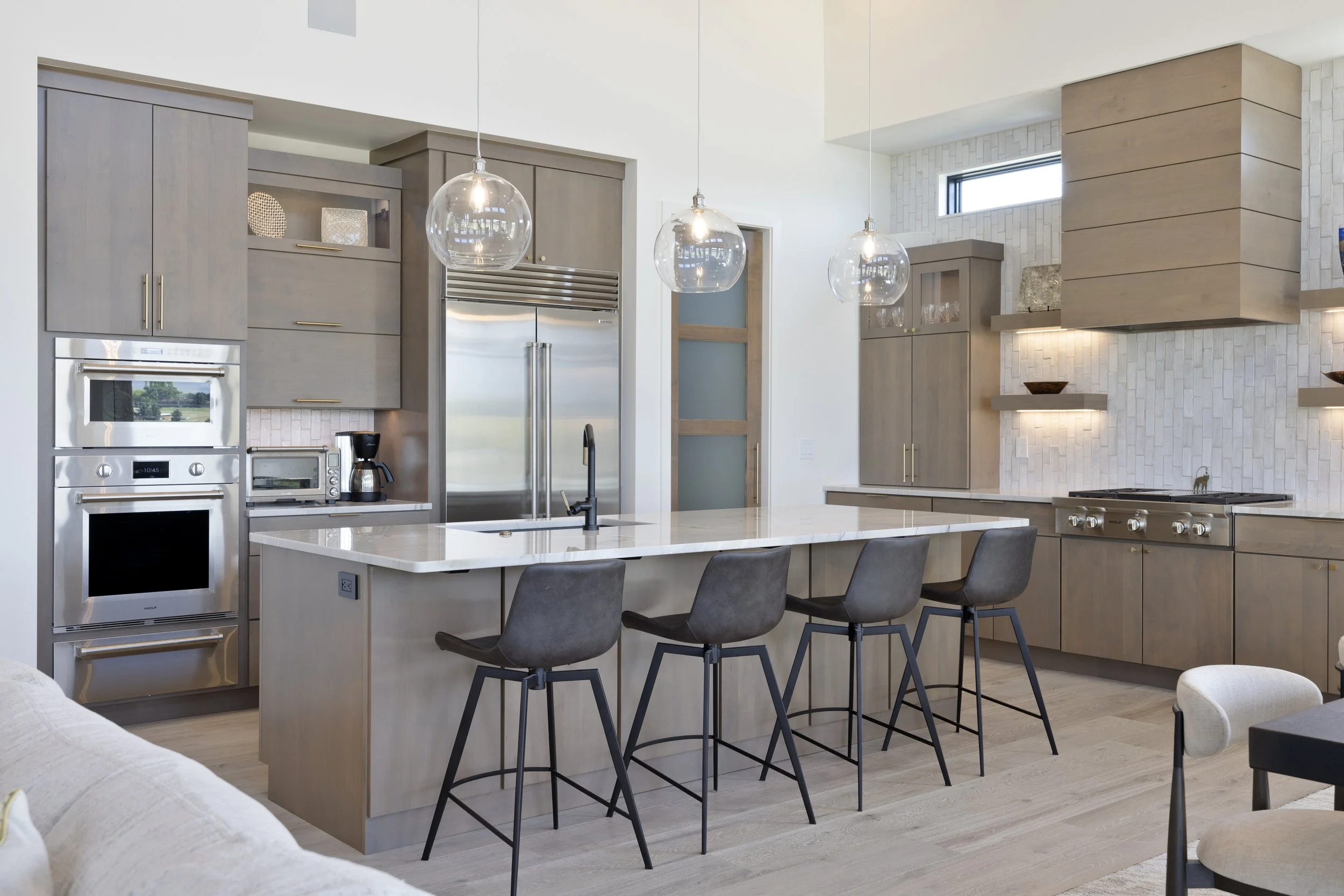 Modern kitchen with gray cabinetry, stainless steel appliances, a white island with a marble countertop, and three hanging glass pendant lights above the island. There are gray barstools around the island and a white brick backsplash behind the stove.