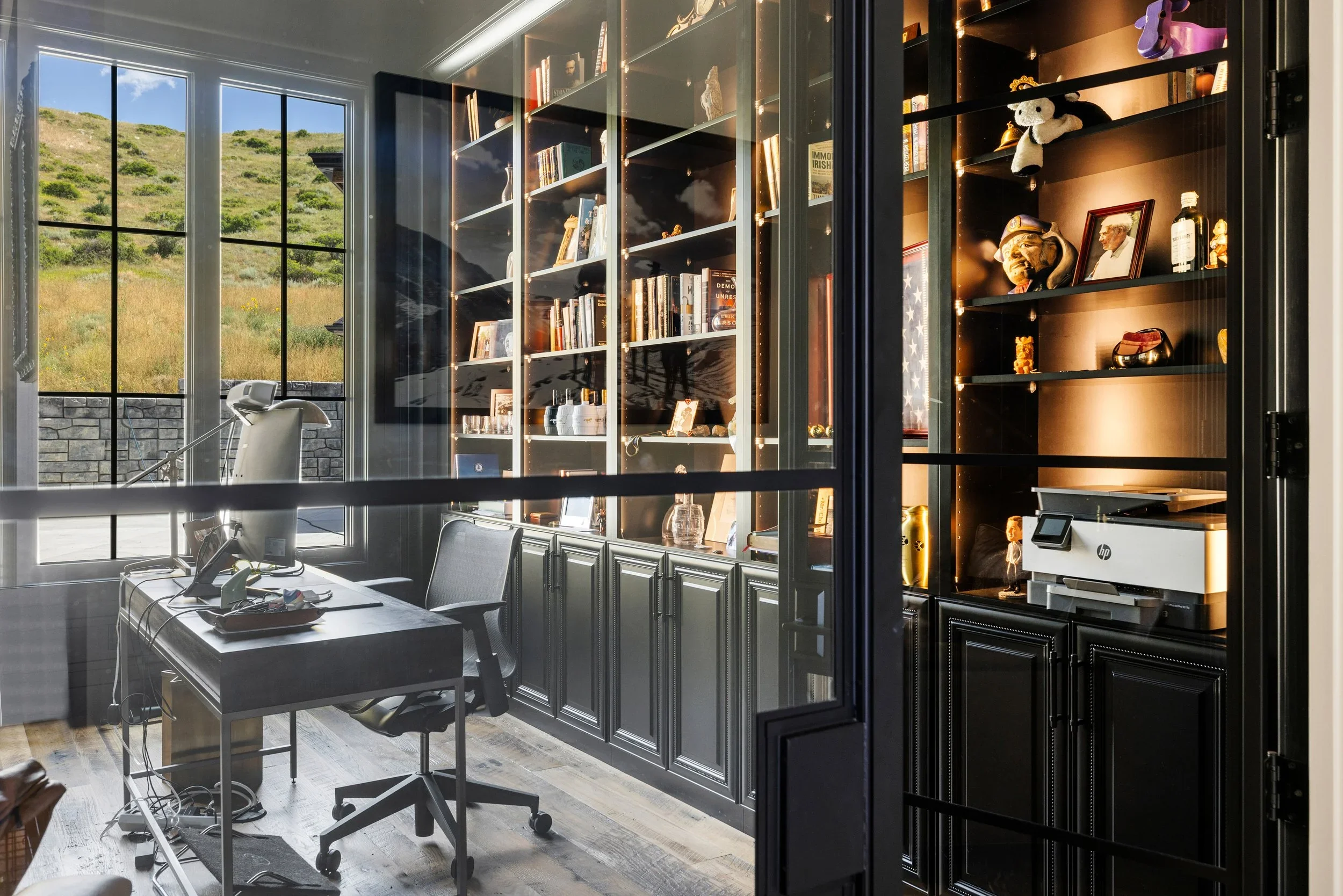 Modern office space with a large black shelving unit filled with books, framed photos, and decorative items next to large windows showing a hillside landscape. A white office chair and a desk with office supplies are in the foreground.