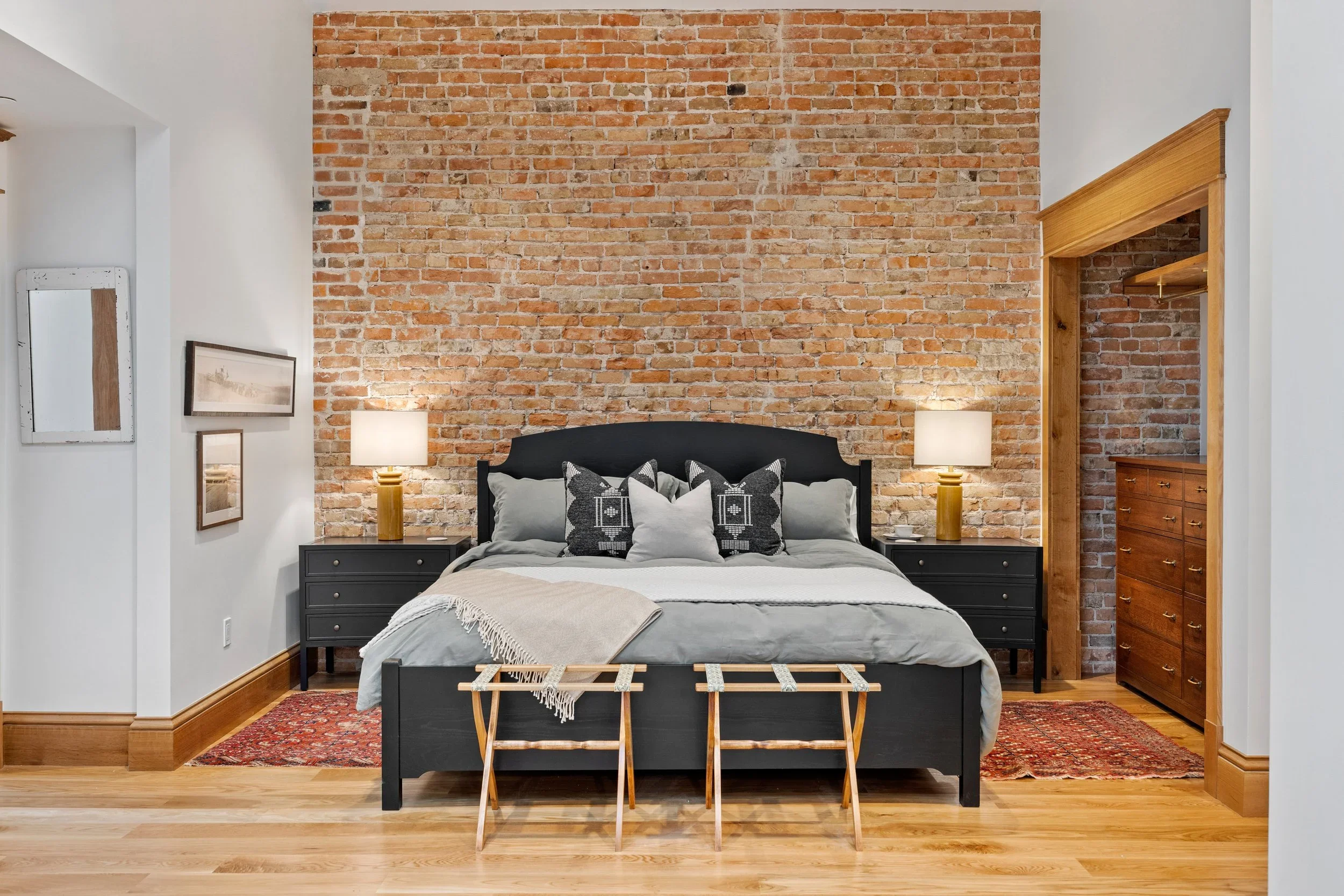 A bedroom with a brick accent wall behind a black bed frame with pillows and a blanket. There are two black nightstands with white lamps, a wooden luggage rack in front of the bed, and a dresser on the right. The room has white walls, wooden trim, and a partially visible closet with a wooden shelf on the right.