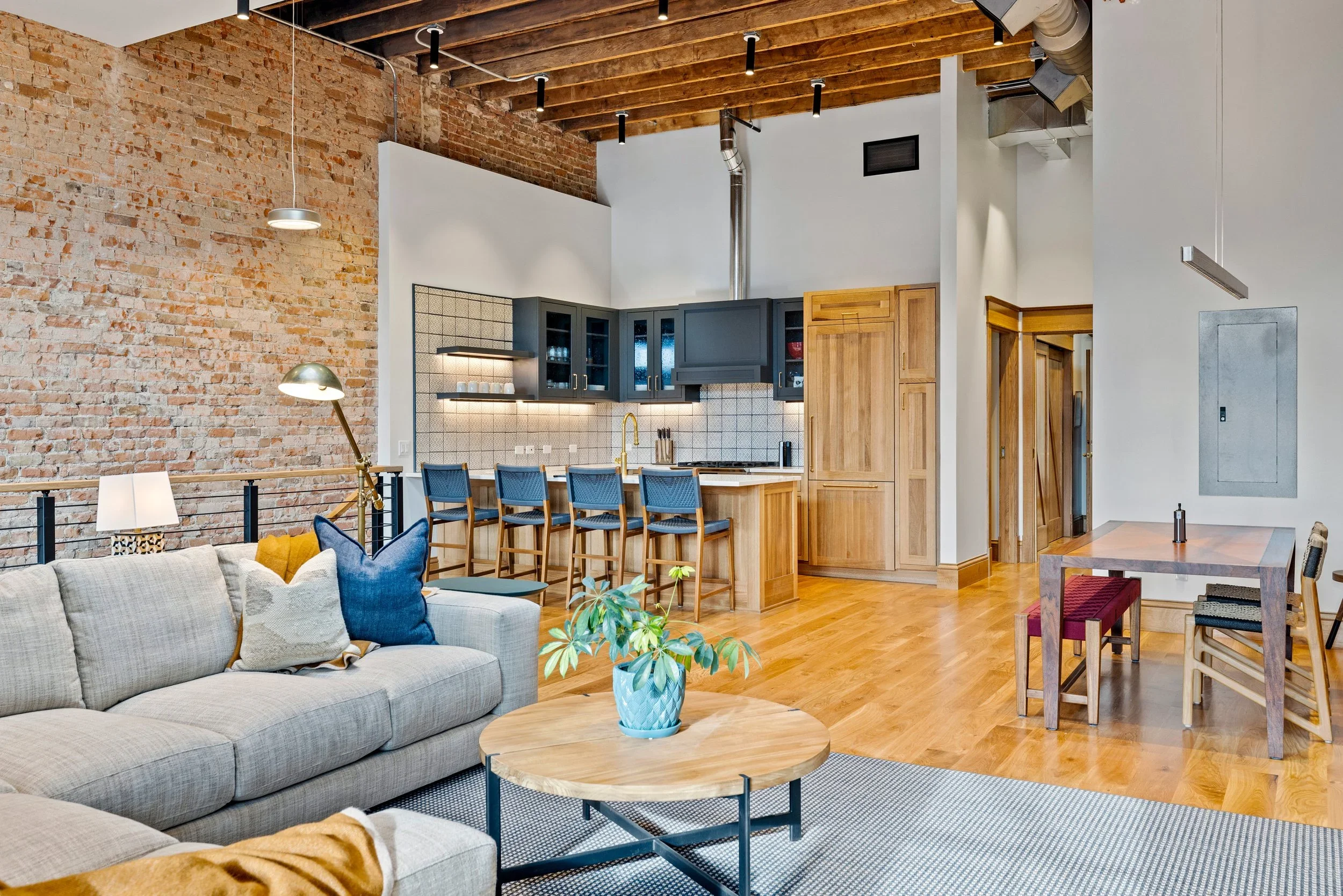 Living room with exposed brick wall, beige sofa with blue pillows, wooden coffee table with plant, open kitchen with dark upper cabinets, wooden lower cabinets, black backsplash, and five bar stools at the kitchen island