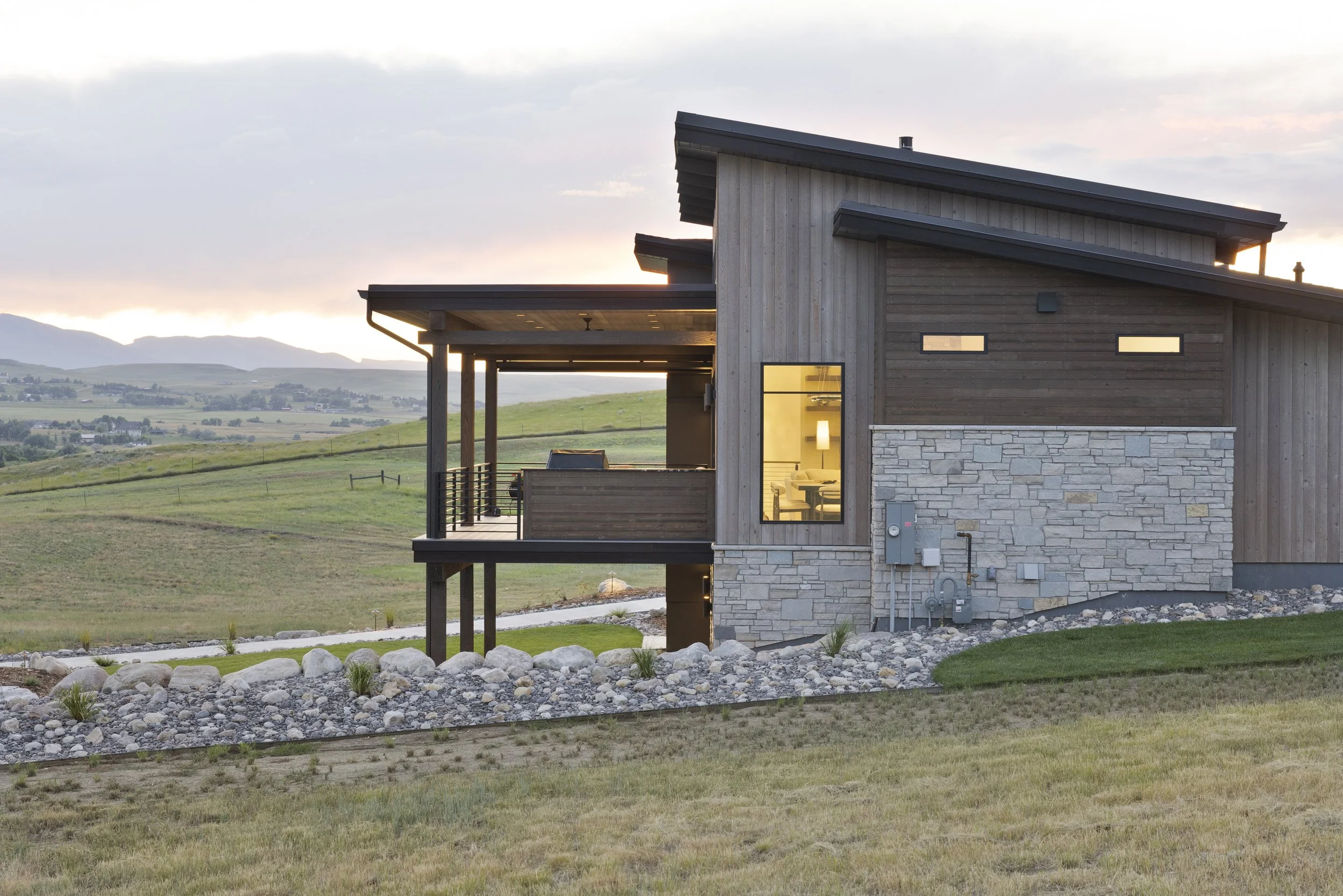 Modern house with wooden and stone exterior in a rural landscape during sunset, featuring a balcony and large windows.