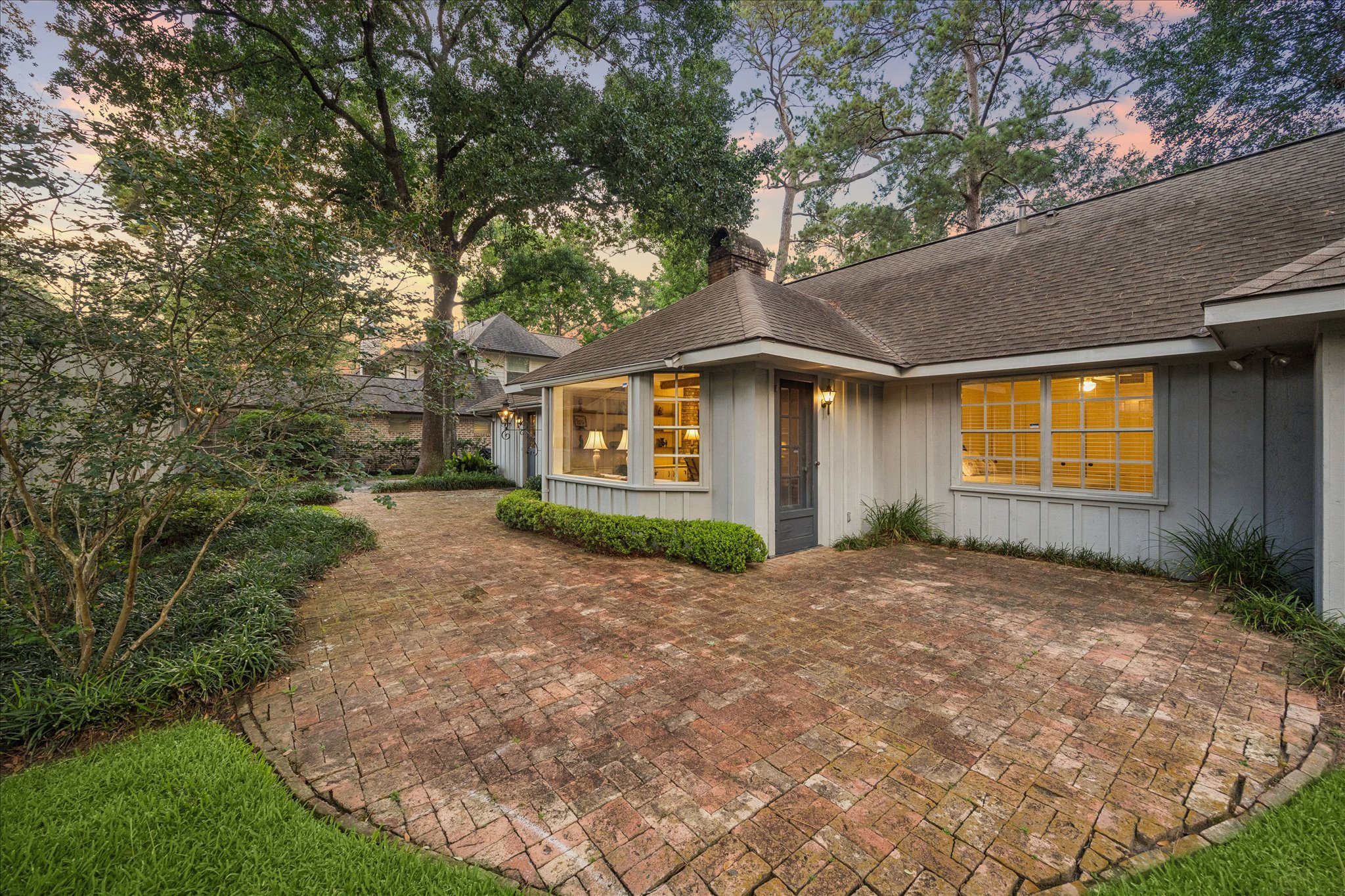 Backyard with brick patio, lush greenery, trees, and a house with illuminated windows and a chimney at sunset.