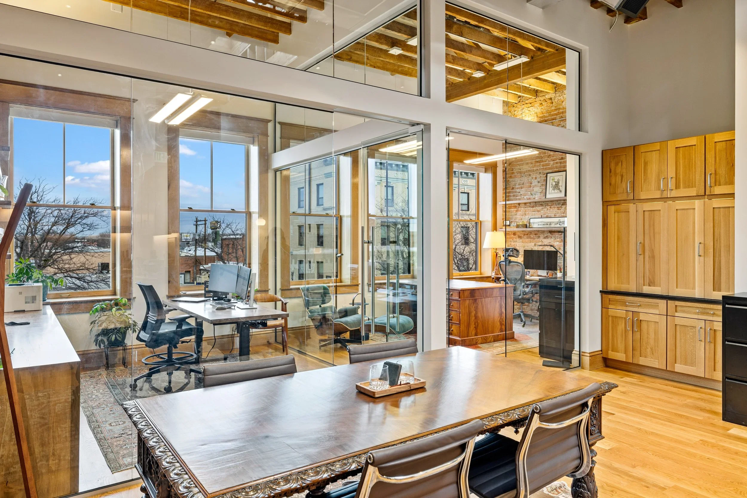 An office space with a large wooden table and ornate chairs in the foreground. Behind, there's a glass room with multiple workstations and computers, large windows showing a cityscape, and wooden ceiling beams. Inside the glass room, there are additional chairs, desks, and a brick wall with a shelf and a framed picture.