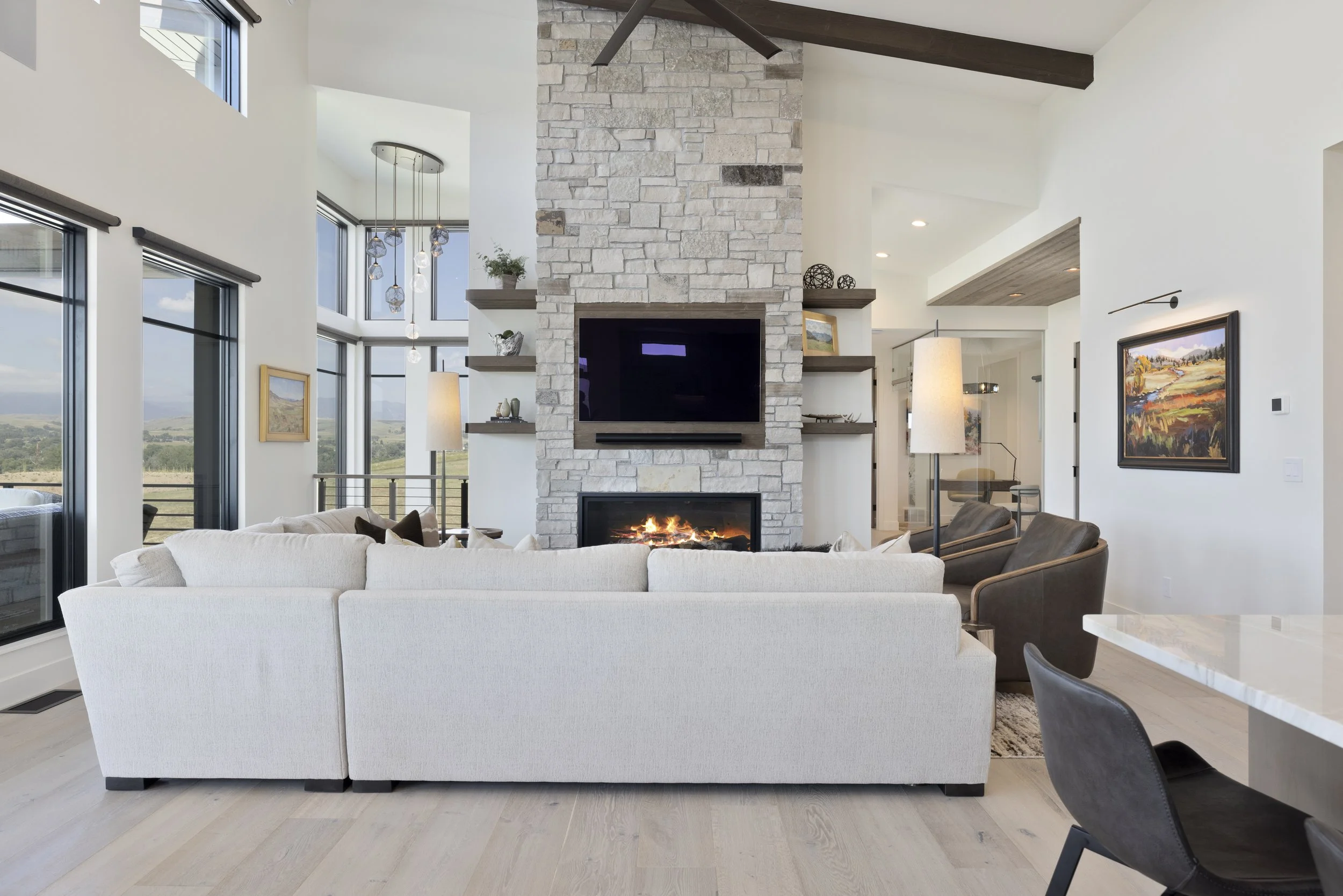Modern living room with a white sectional sofa, a stone fireplace with a TV above, and large windows showing a scenic view outside.