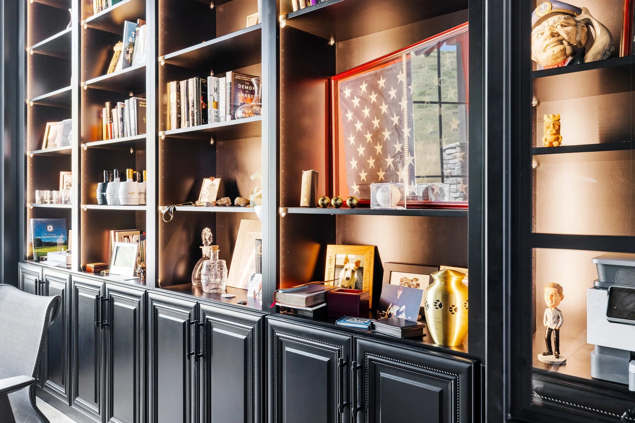 Black bookshelf with books, framed pictures, decorative items, American flag artwork, and stuffed animals in a room with a gray chair nearby.