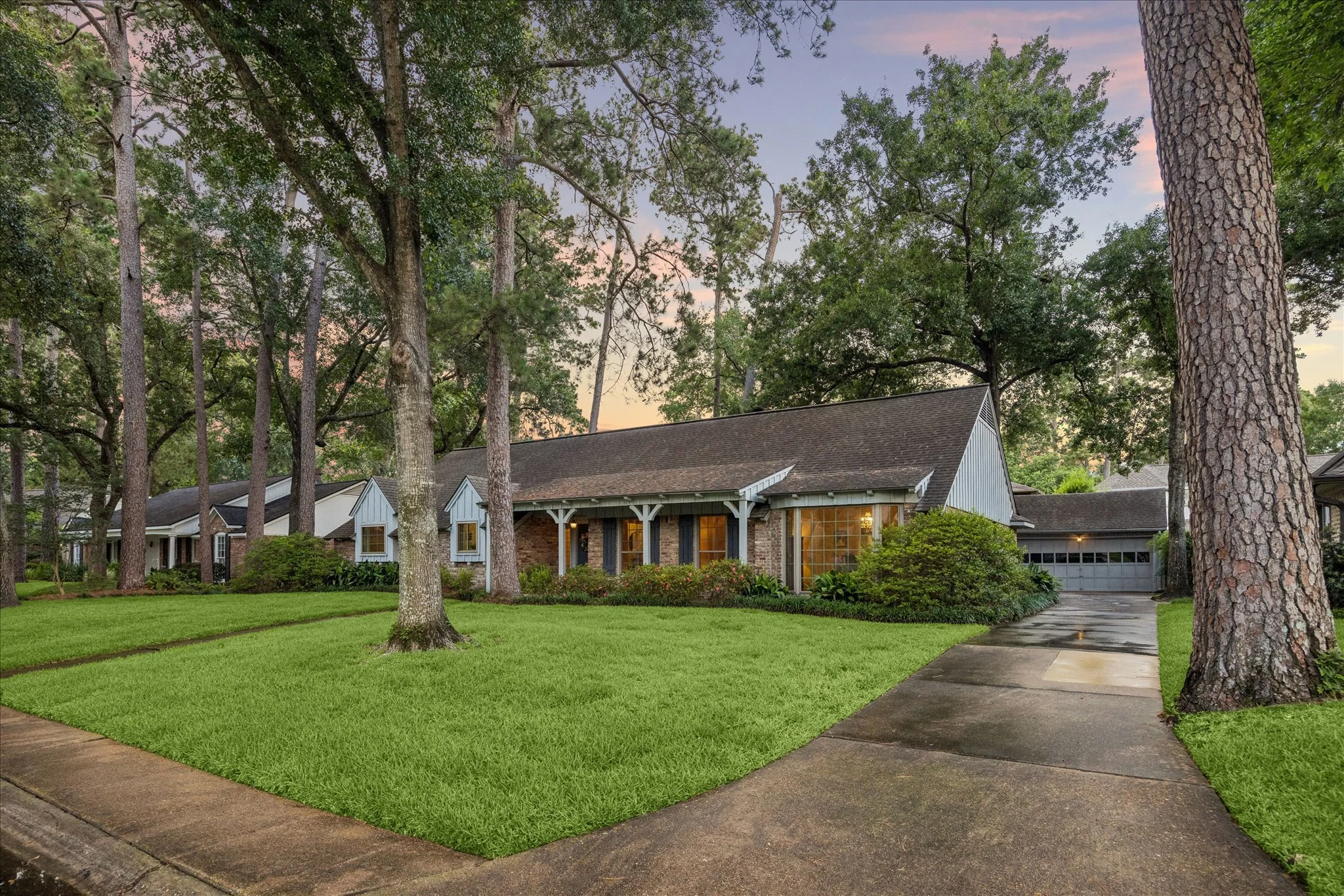 A suburban house with a brick and white siding exterior, large windows, and a front porch with support columns. Surrounding the house are tall trees, green lawn, and a sidewalk leading to the driveway, during dusk.