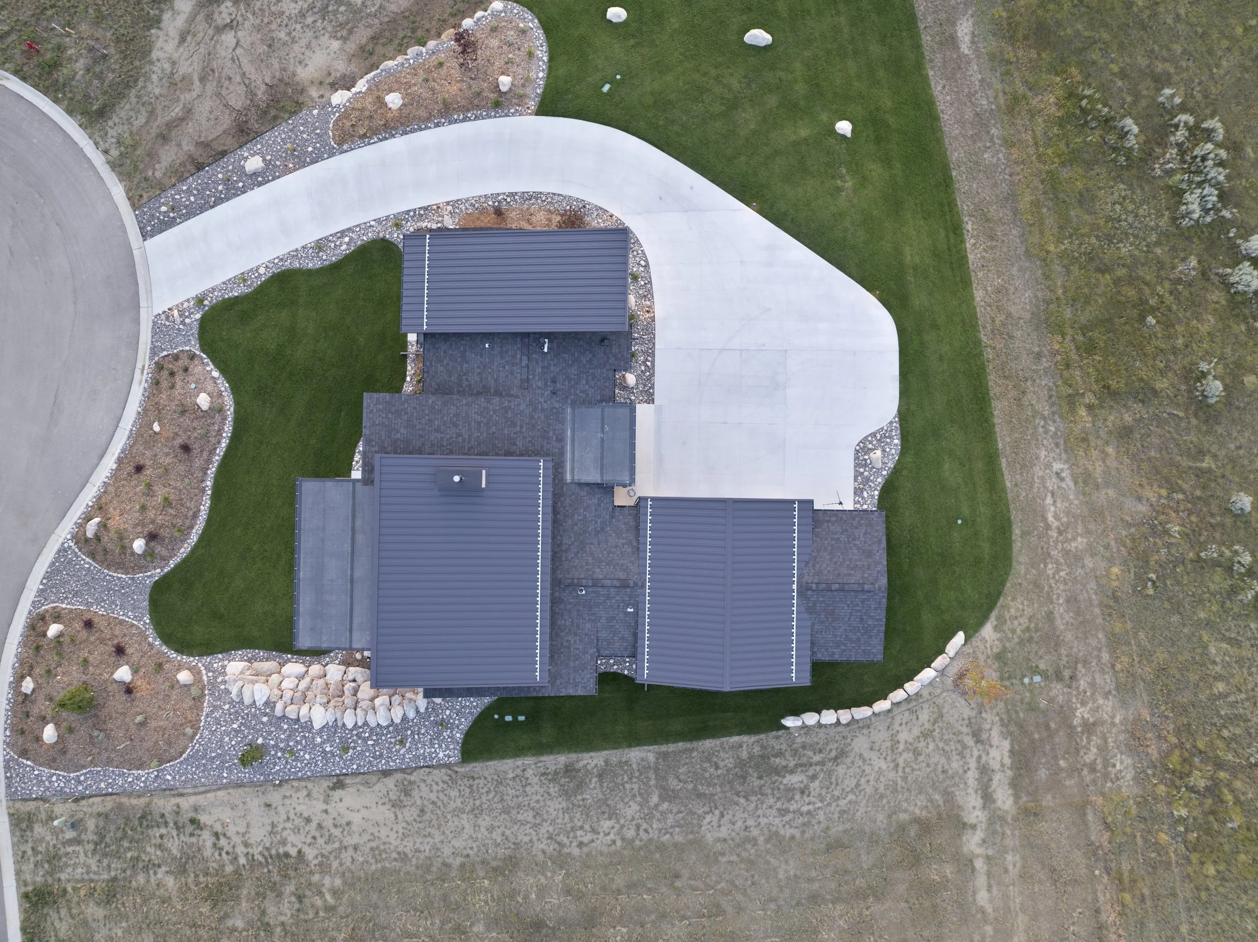 An aerial view of a modern house with a gray roof, surrounded by a manicured lawn, a winding concrete pathway, landscaped garden beds with rocks and plants, and undeveloped land with sparse vegetation.