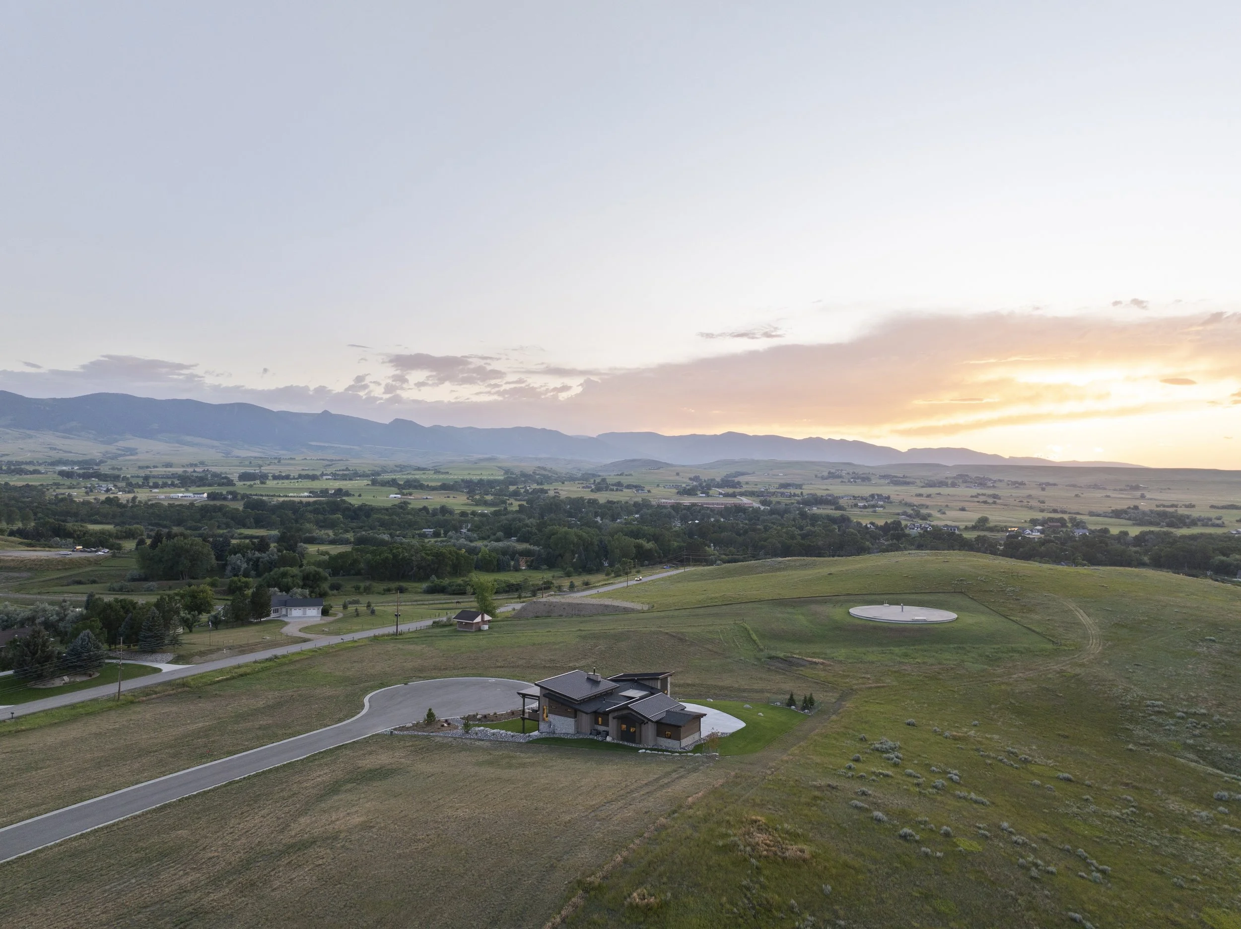 A scenic landscape view at sunset with hills in the distance, a green valley, and a few houses, seen from an elevated position.